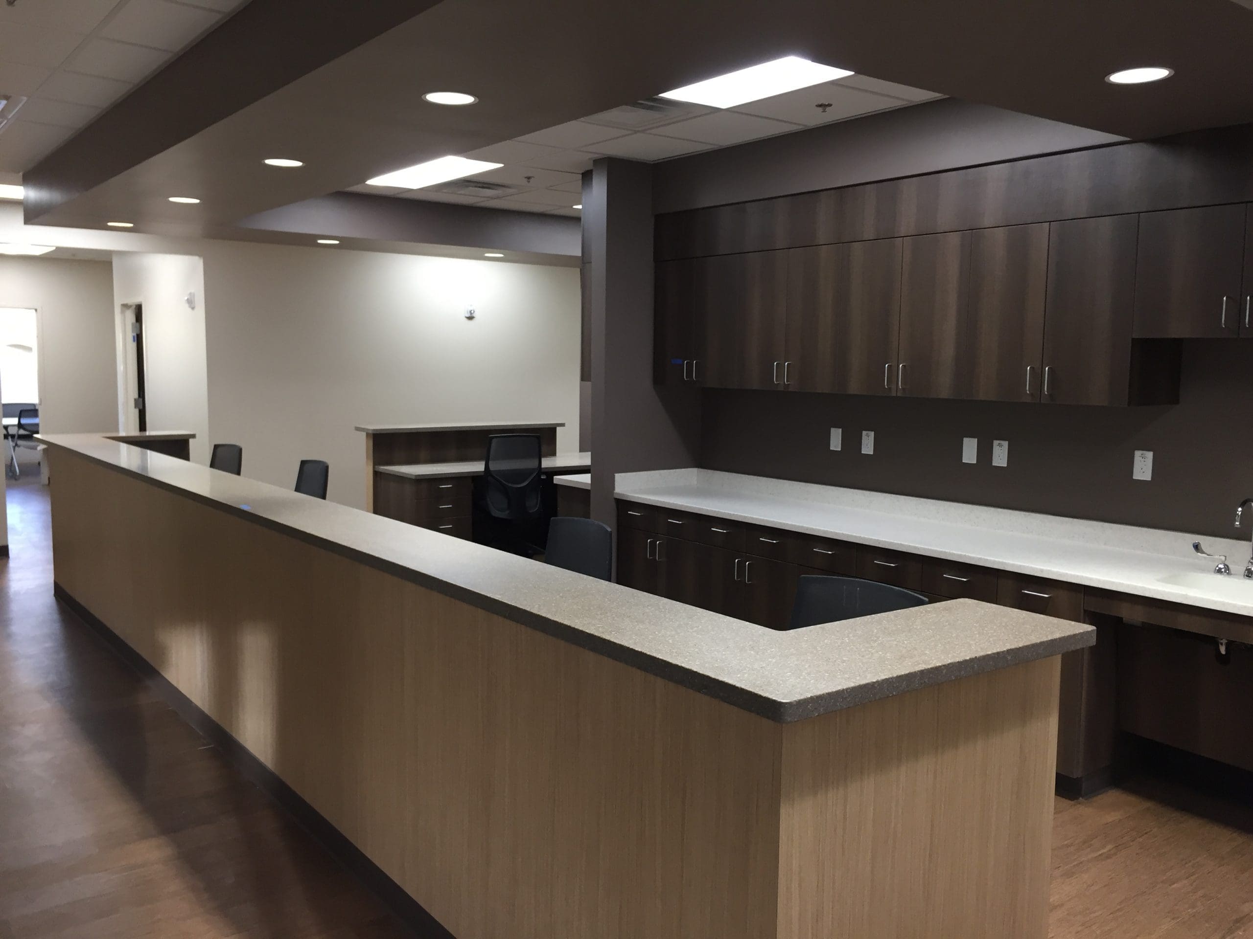 A modern reception or nurse station with wood cabinetry, white countertops, several chairs, and overhead lighting in a medical or office facility.