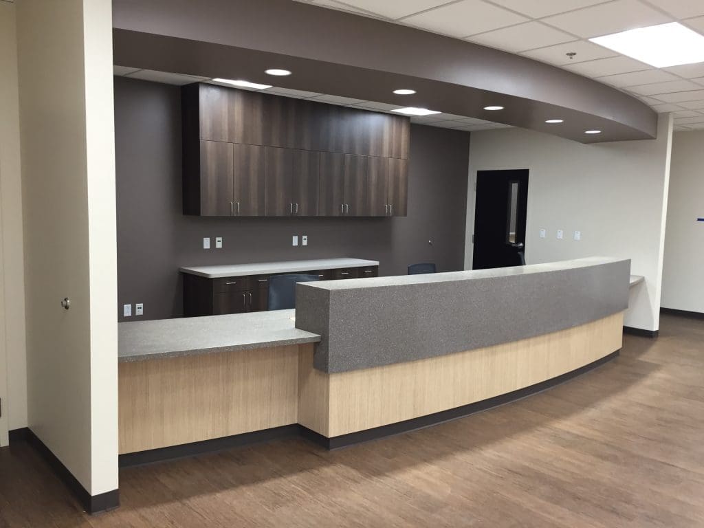 A modern, empty reception desk with wood and grey finishes, set against a neutral-toned wall with cabinets and overhead lighting.