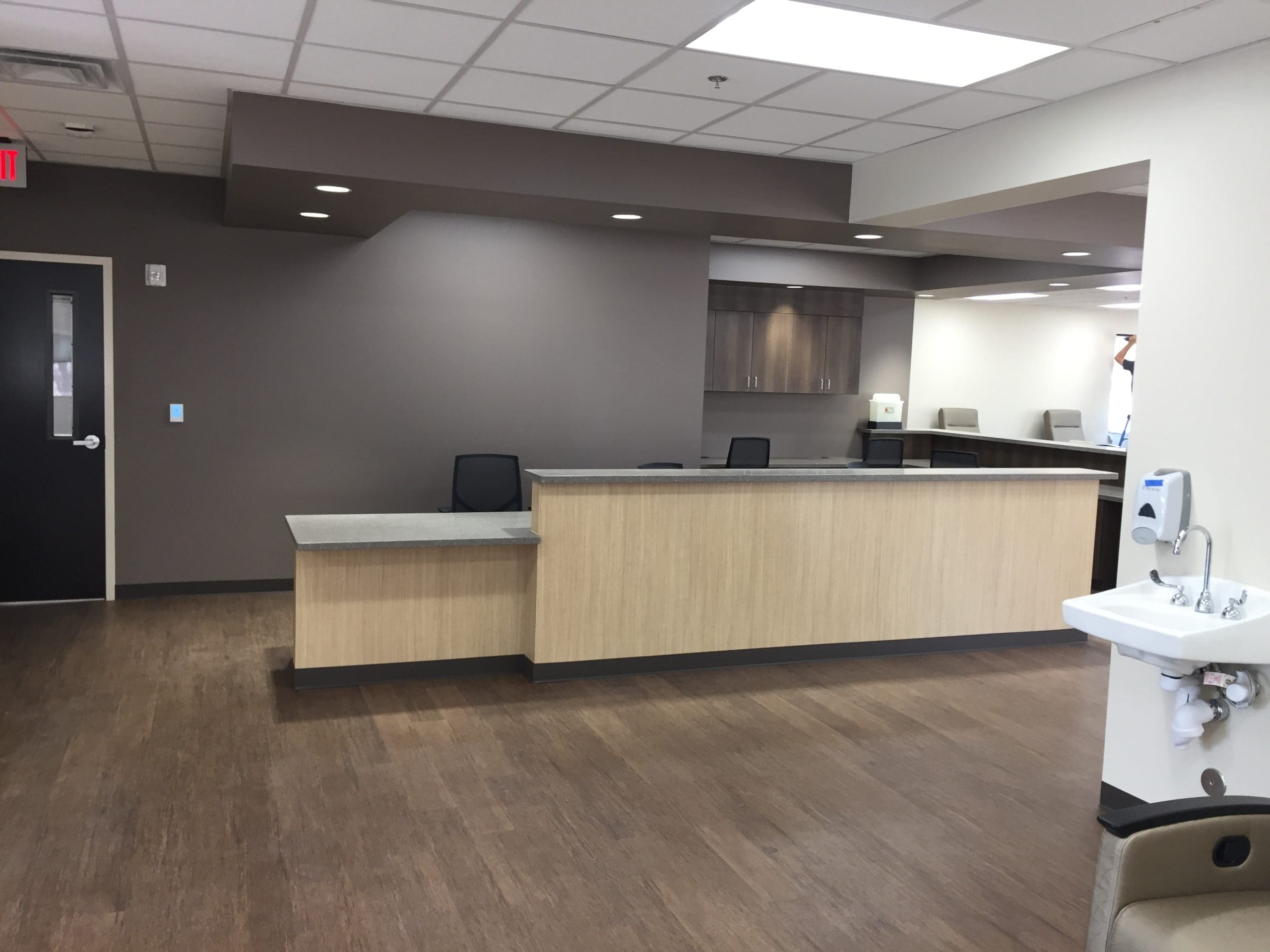 A modern, empty medical office reception area with a wooden front desk, brown flooring, chairs, and a wall-mounted sink in the foreground.