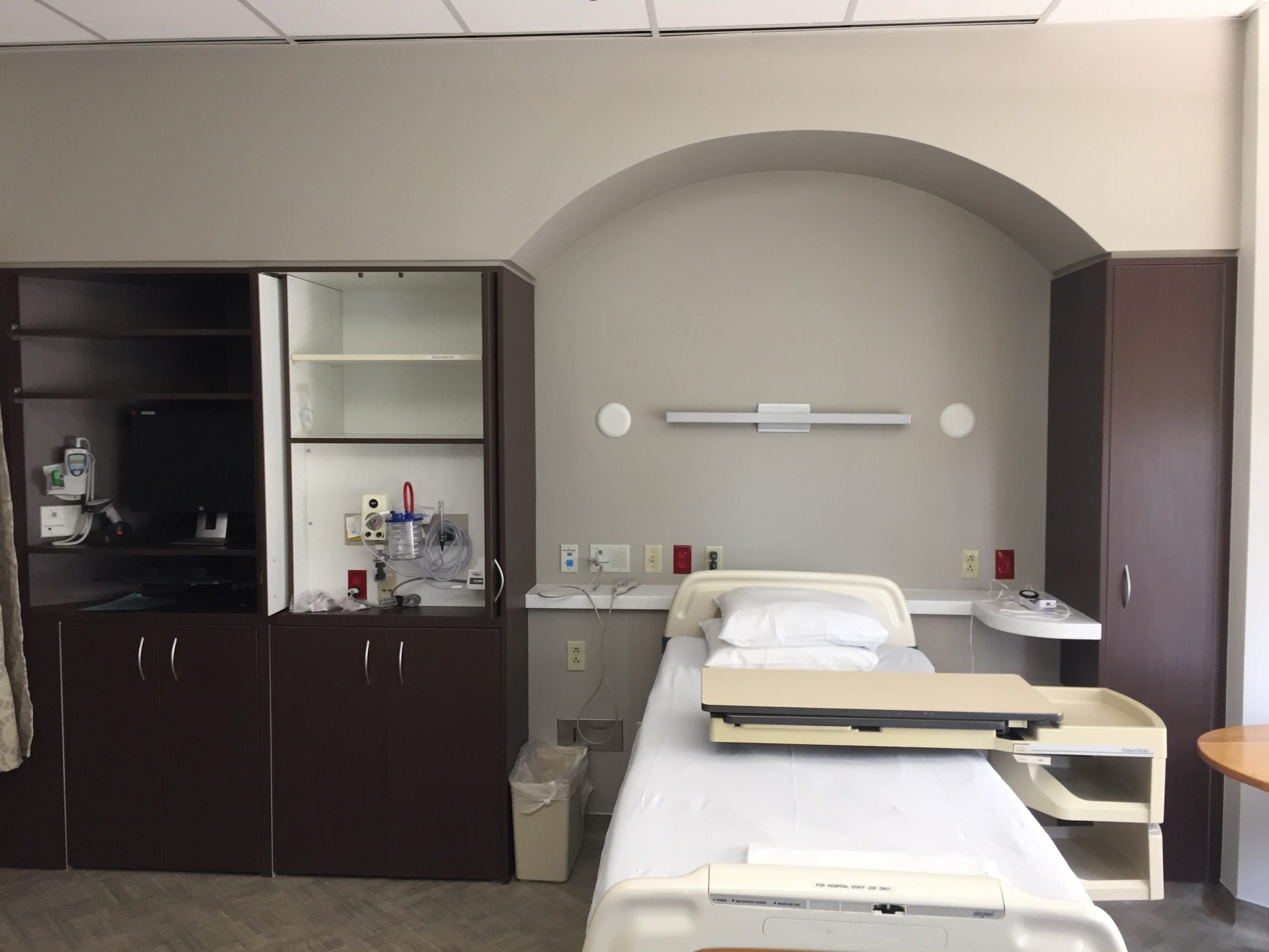 A single hospital bed with white linens and a bedside tray, surrounded by built-in cabinets, medical equipment, and various wall outlets in a clean patient room.