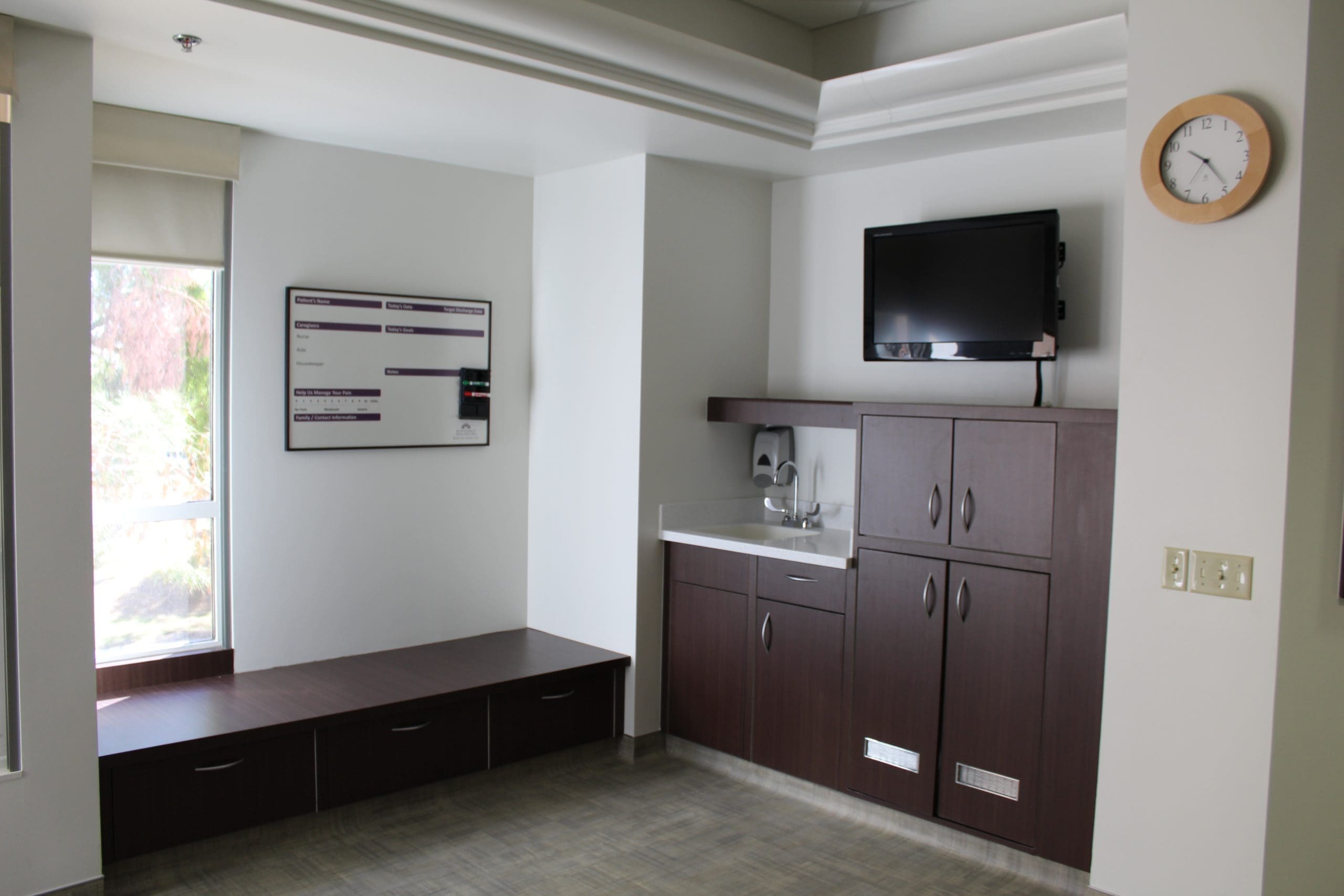 A hospital or clinic room corner with a wall clock, TV, whiteboard, cabinets, countertop with sink, and large window.