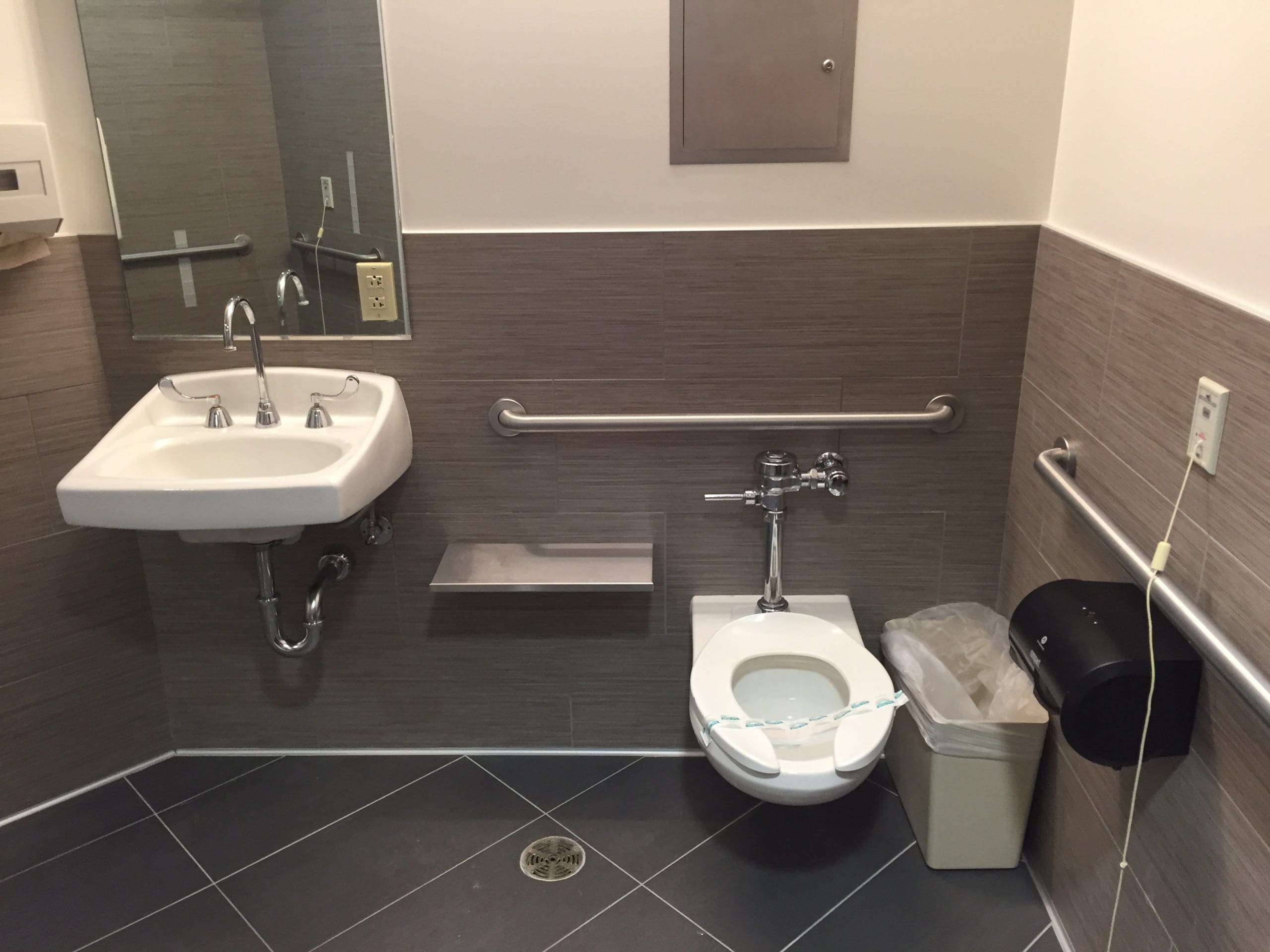 Accessible restroom with a wall-mounted sink, toilet with grab bars, waste bin, and toilet paper dispenser; toilet has a strip indicating it has been sanitized.