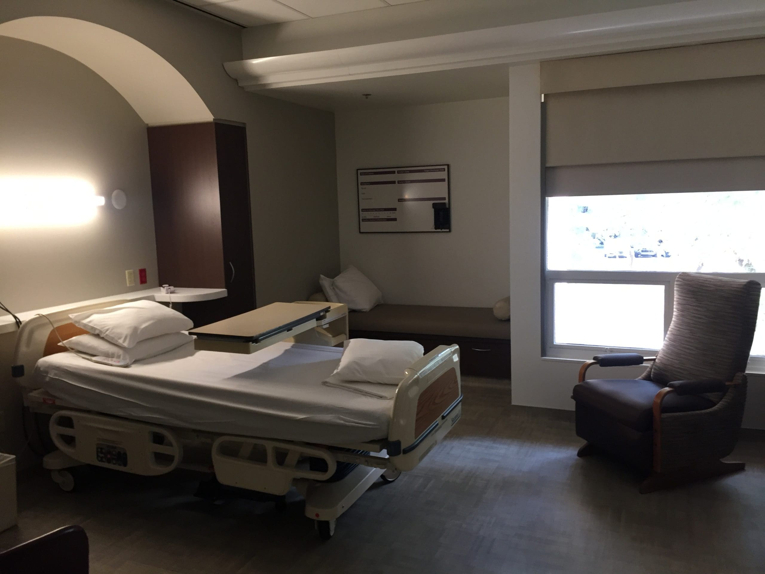 A hospital room with an empty bed, a tray table, a cushioned bench, a chair, and a window with the shade partially drawn.