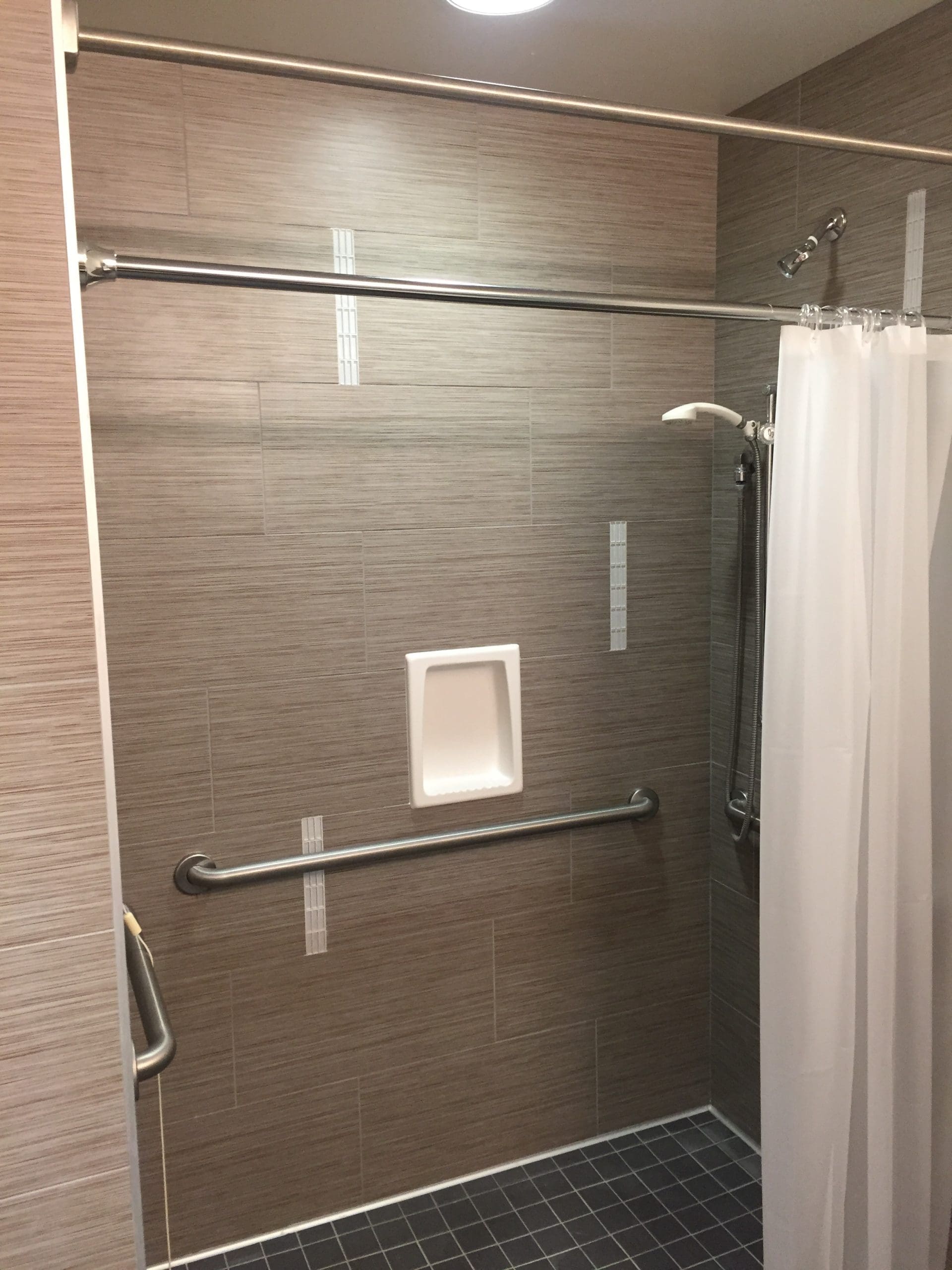 Accessible shower with gray tile, a white shower curtain, two metal grab bars, built-in soap niche, and a handheld showerhead.