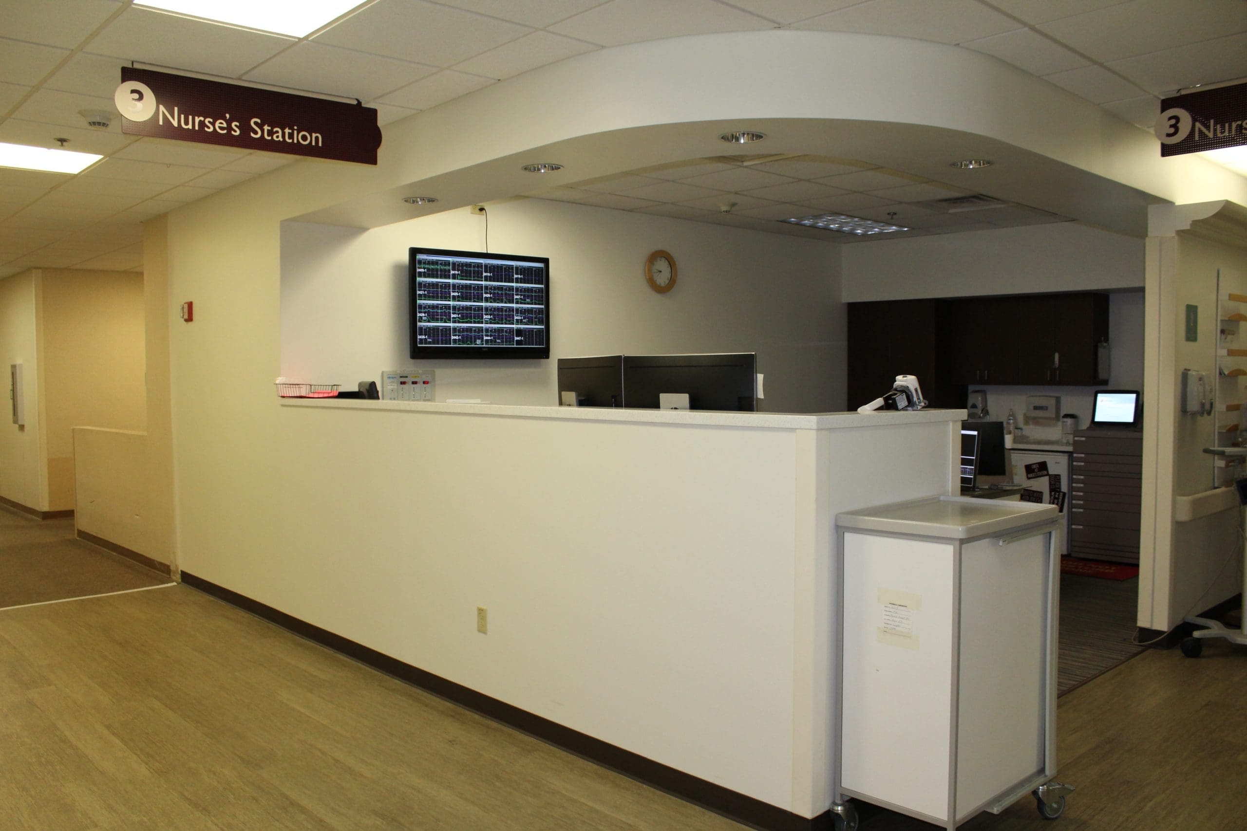 A hospital nurse’s station with monitors on the wall, computer desks, and medical equipment in a clean, well-lit hallway.