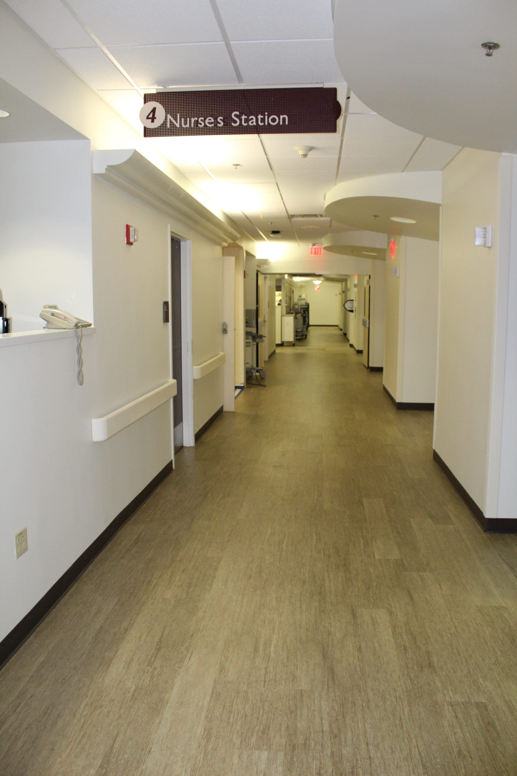 A hospital corridor with beige floors and white walls, featuring a sign overhead that reads "Nurses' Station" and medical equipment visible in the distance.
