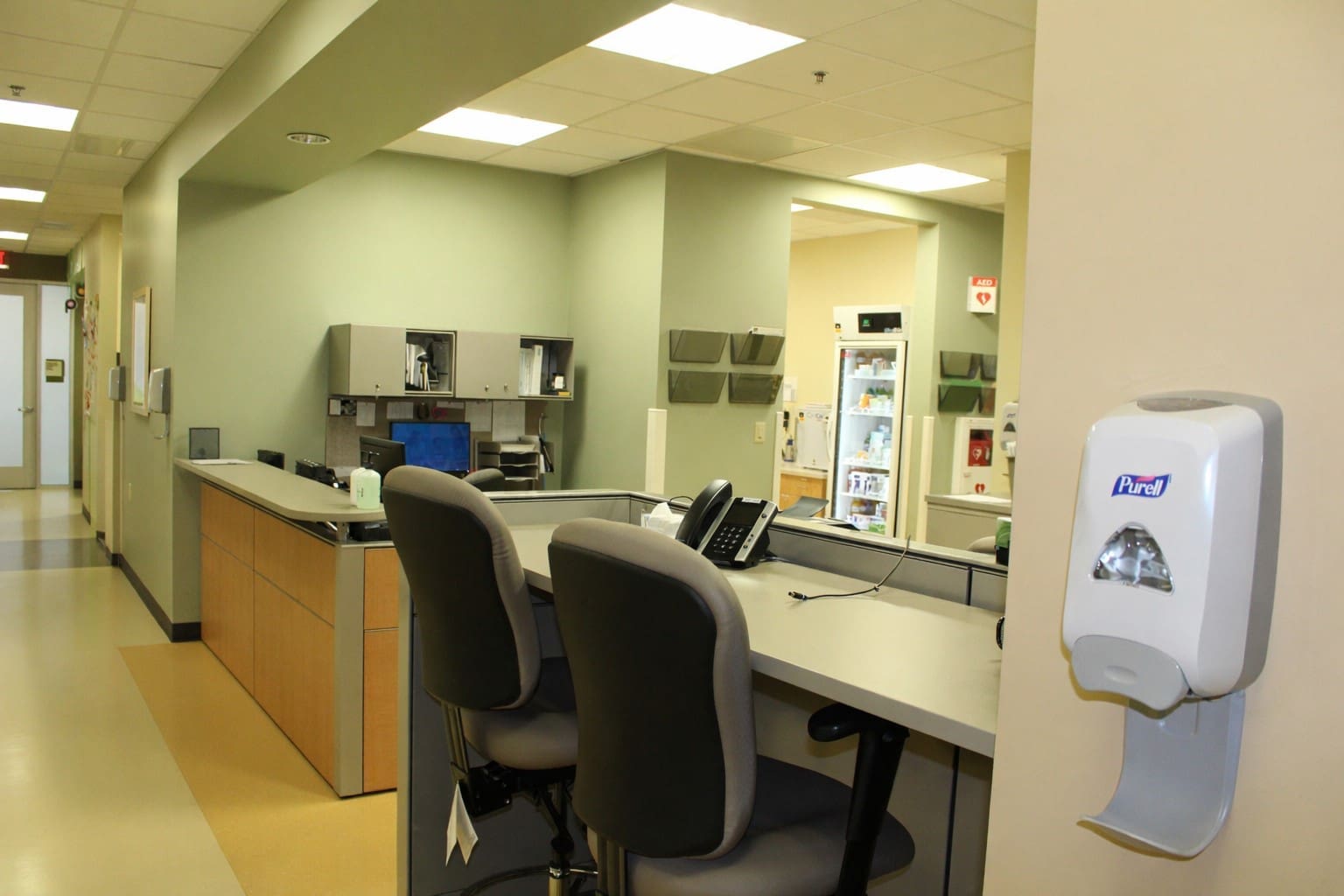 A hospital or clinic nurses' station with empty chairs, a desk with office equipment, cabinets, a computer, and a wall-mounted hand sanitizer dispenser in the foreground.