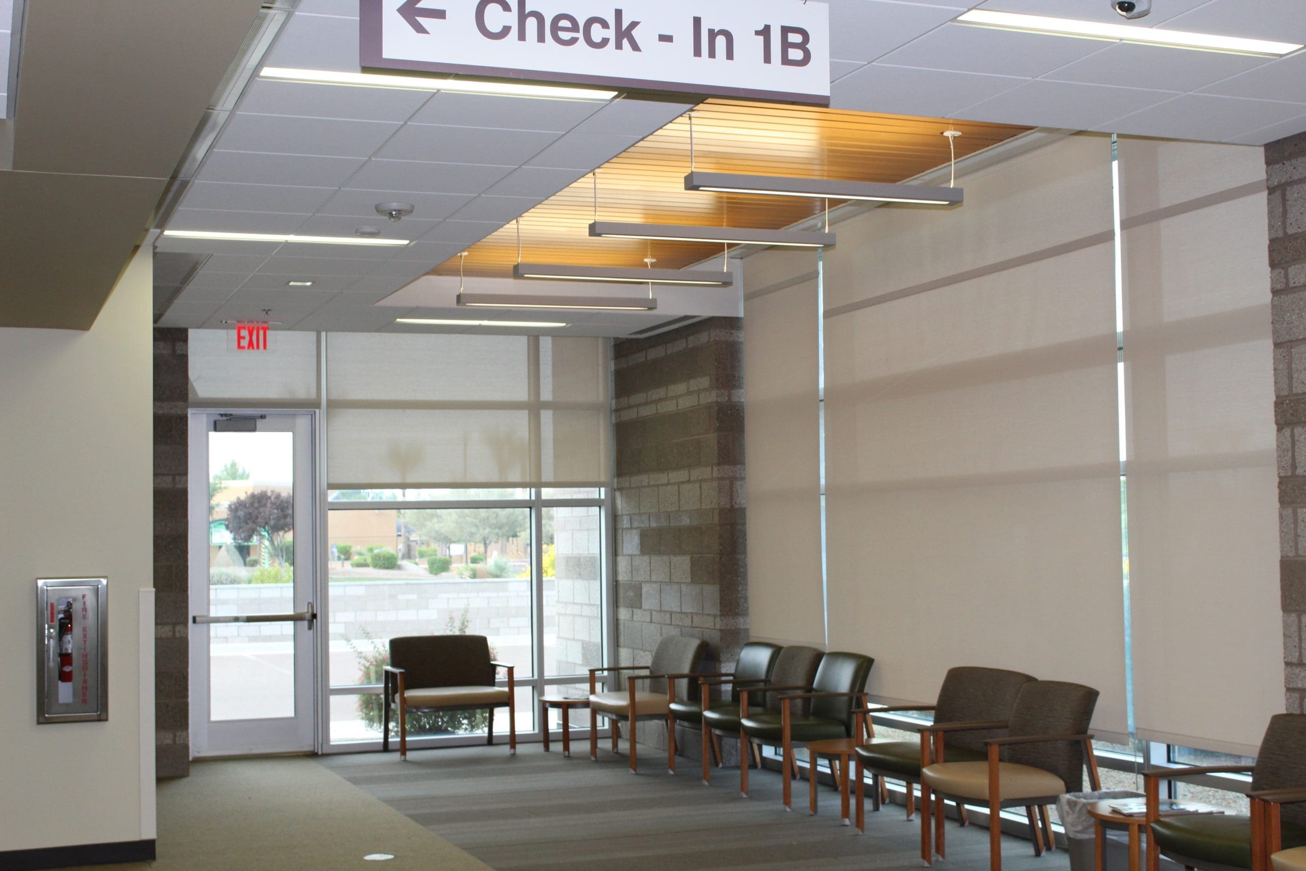 A waiting area with empty chairs, large windows with blinds, and a sign overhead reading "Check-In 1B.