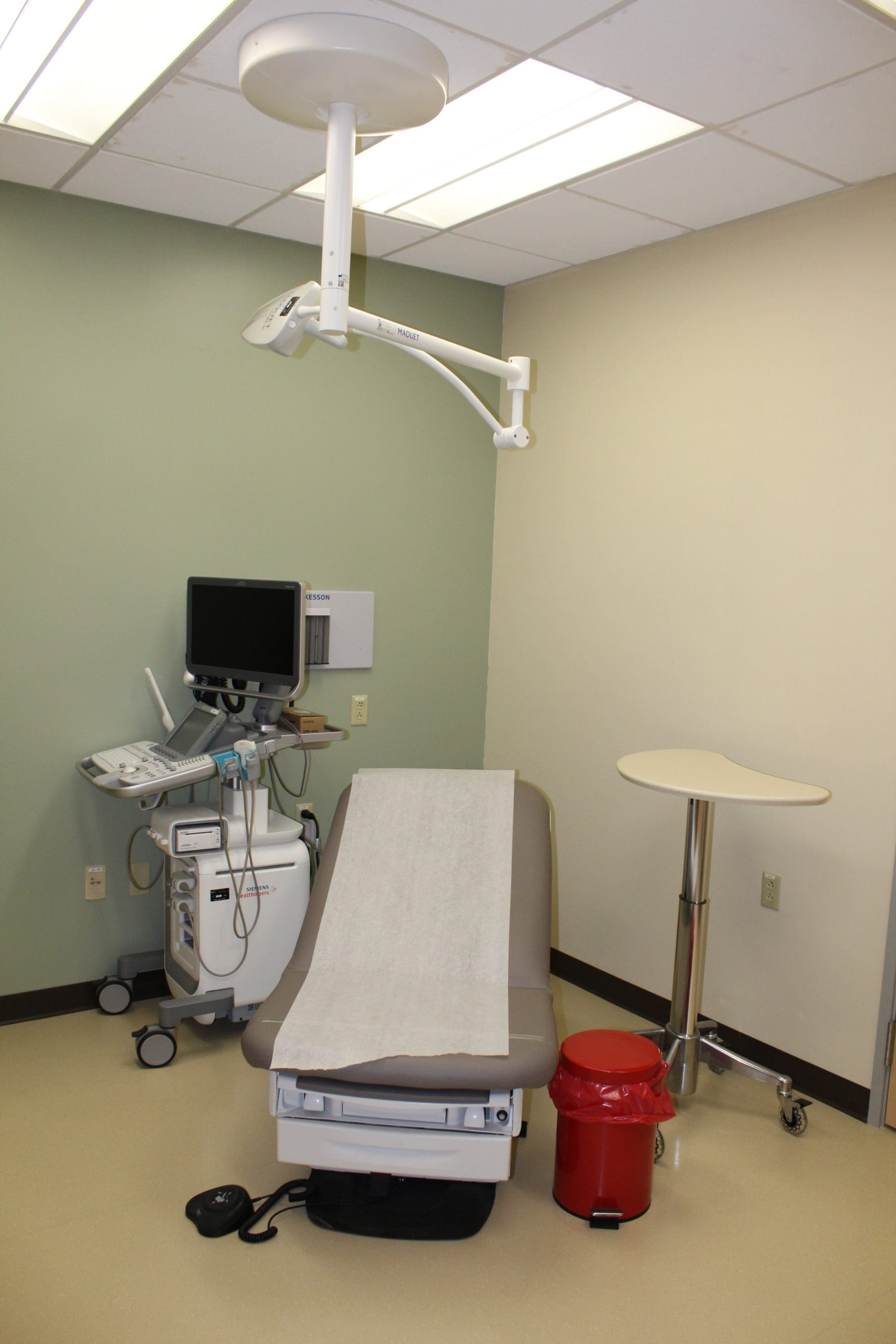 A medical exam room with an examination table, ultrasound machine, red biohazard bin, small round table, and overhead examination light.