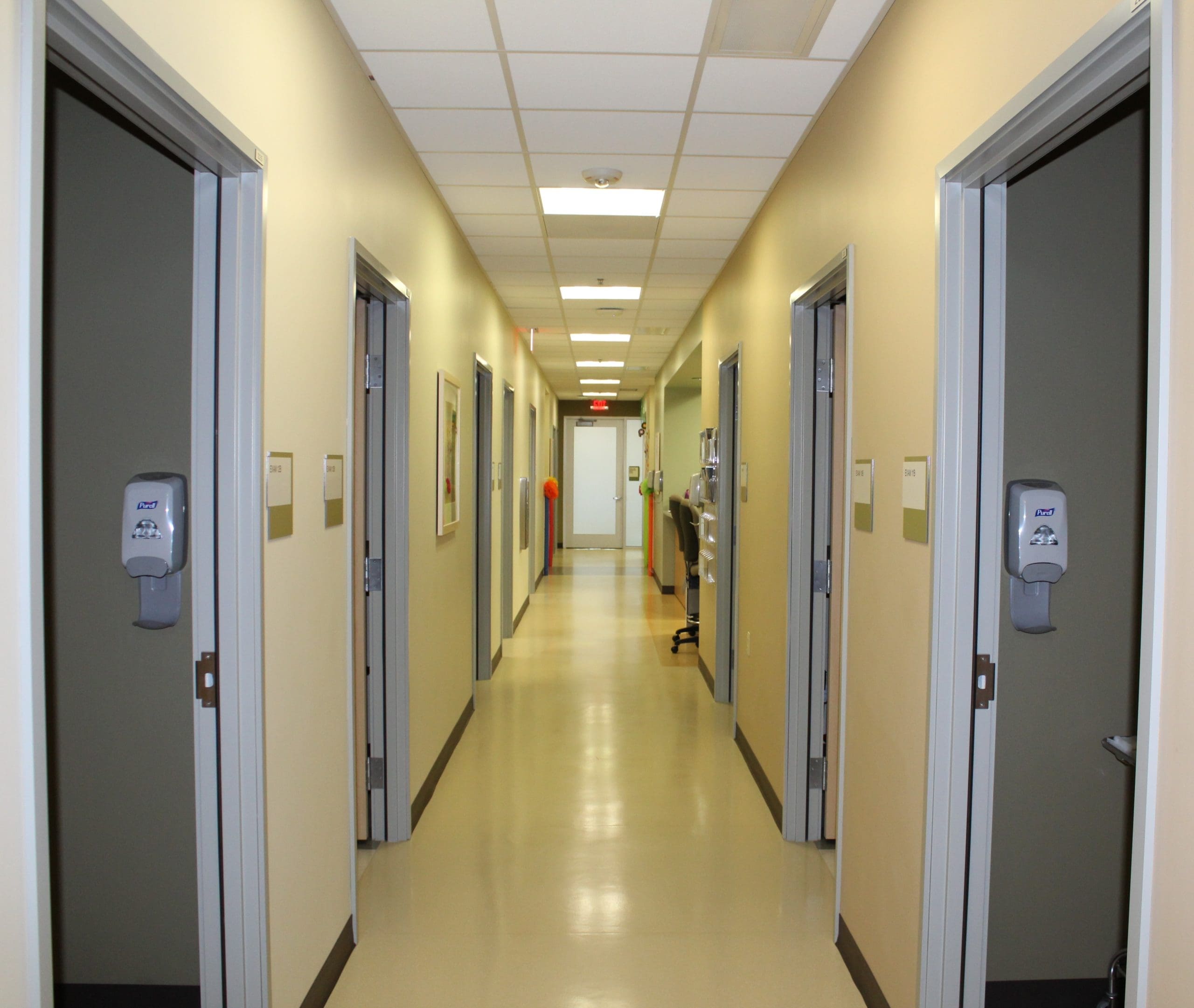 A brightly lit hallway in a medical facility with several open doors and wall-mounted hand sanitizer dispensers.