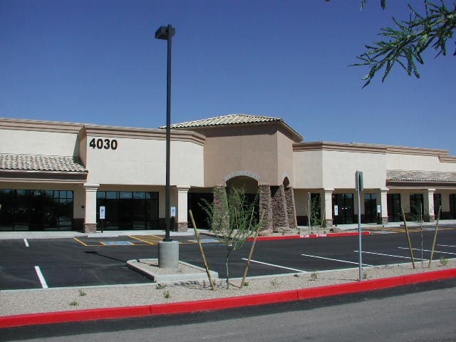 A beige commercial building with the number 4030, empty parking spaces, and a red curb in front under a clear blue sky.