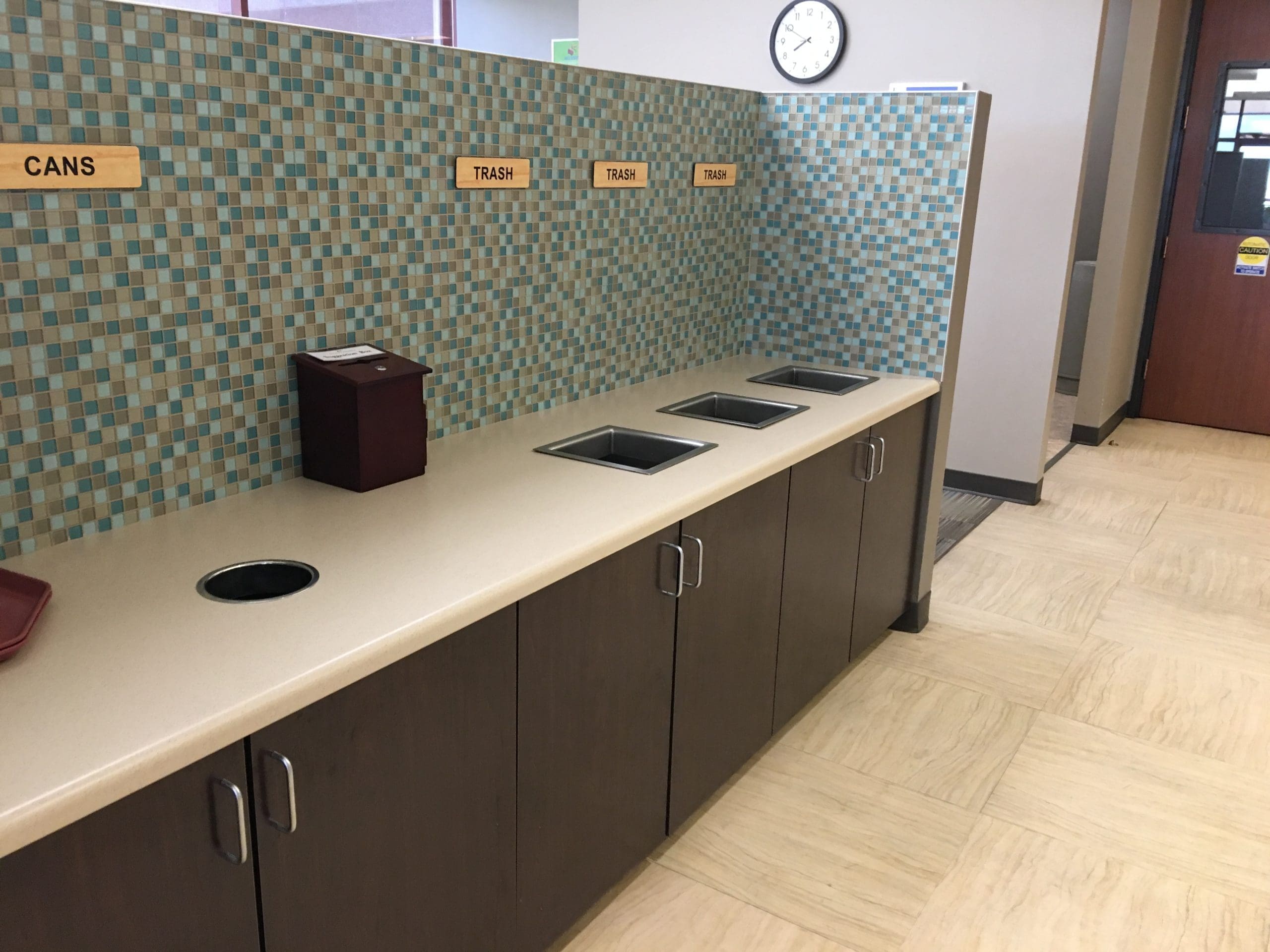 Countertop with labeled waste openings for cans and trash, set against a tiled wall inside a clean indoor facility.