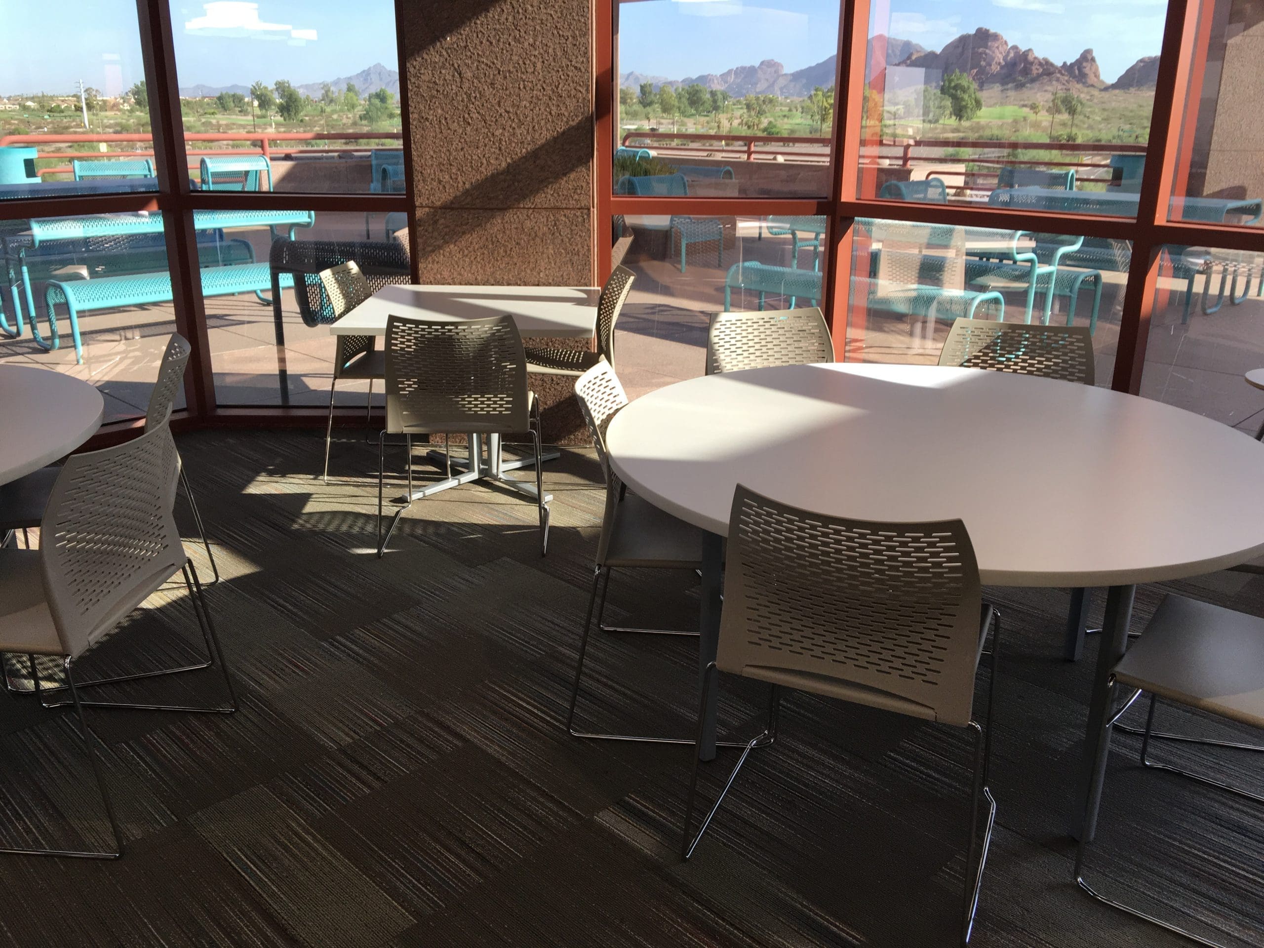 Round tables and gray chairs are arranged in a sunlit indoor seating area with large windows overlooking an outdoor patio and distant mountains.