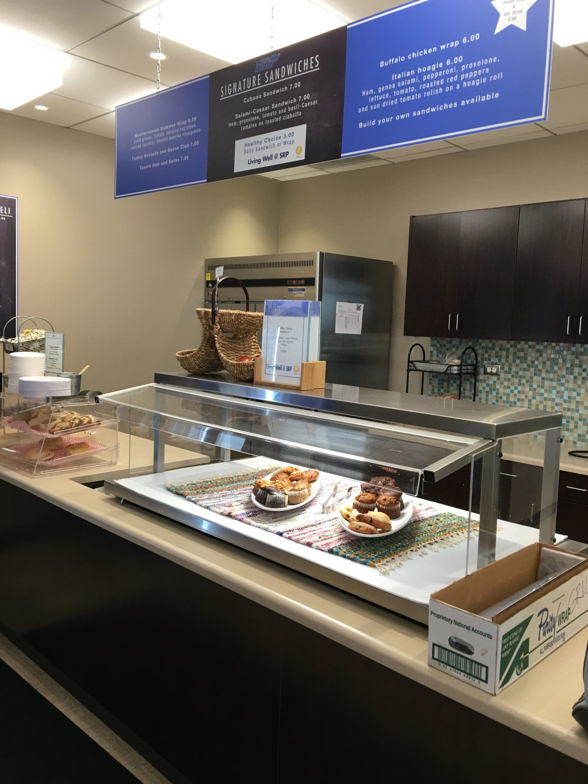 A cafeteria counter displays several pastries on trays under a glass case, with menus hanging overhead and a tiled backsplash in the background.