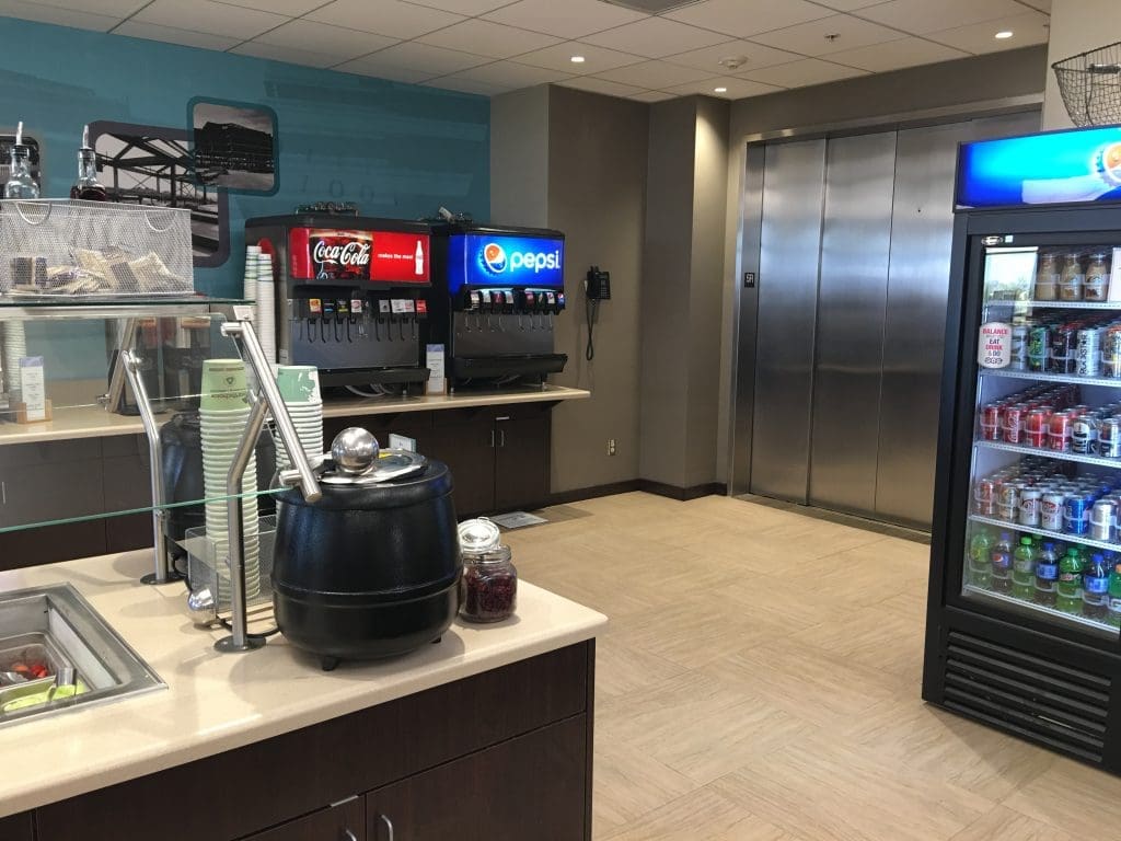 Cafeteria area with a soup station, soda fountain machines, a refrigerator stocked with drinks, and an elevator in the background.