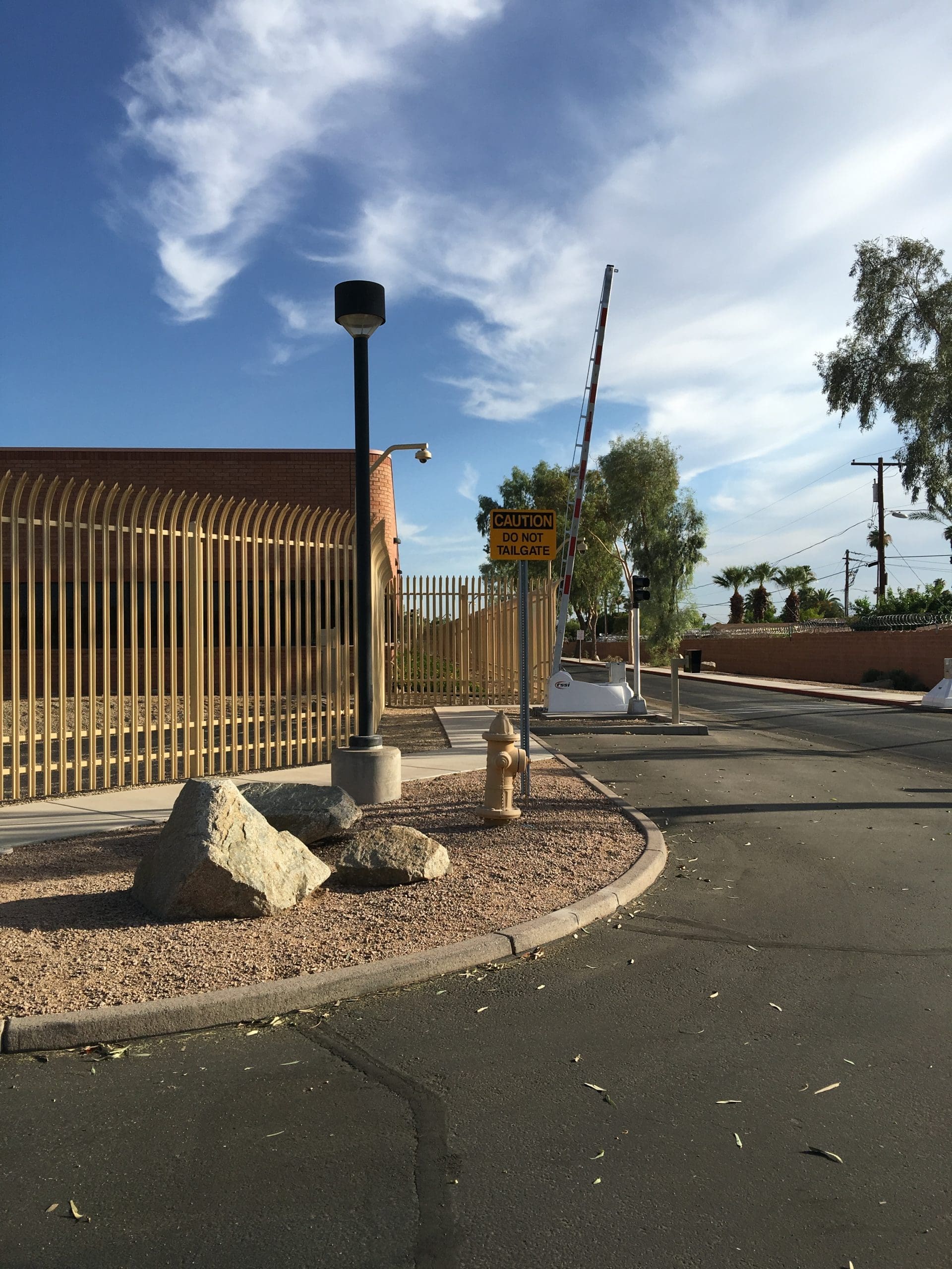 A gated driveway entrance with a caution sign, security camera, barrier arm, fire hydrant, and large rocks near the curb under a clear sky.