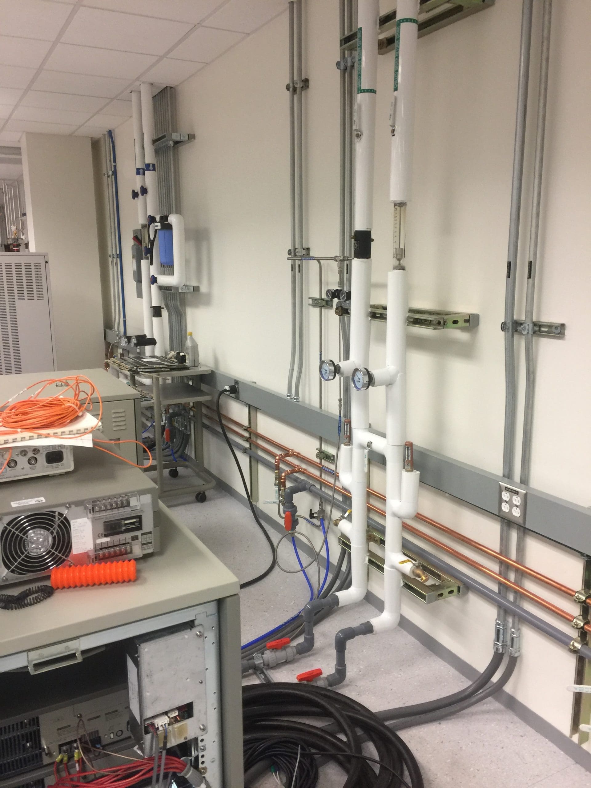 A laboratory room with exposed pipes, cables, gauges, and electronic equipment placed on tables and connected to the wall.