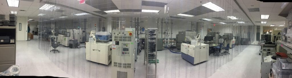 Panoramic view of a cleanroom laboratory with various industrial machines, equipment, and computers behind transparent plastic strip curtains.
