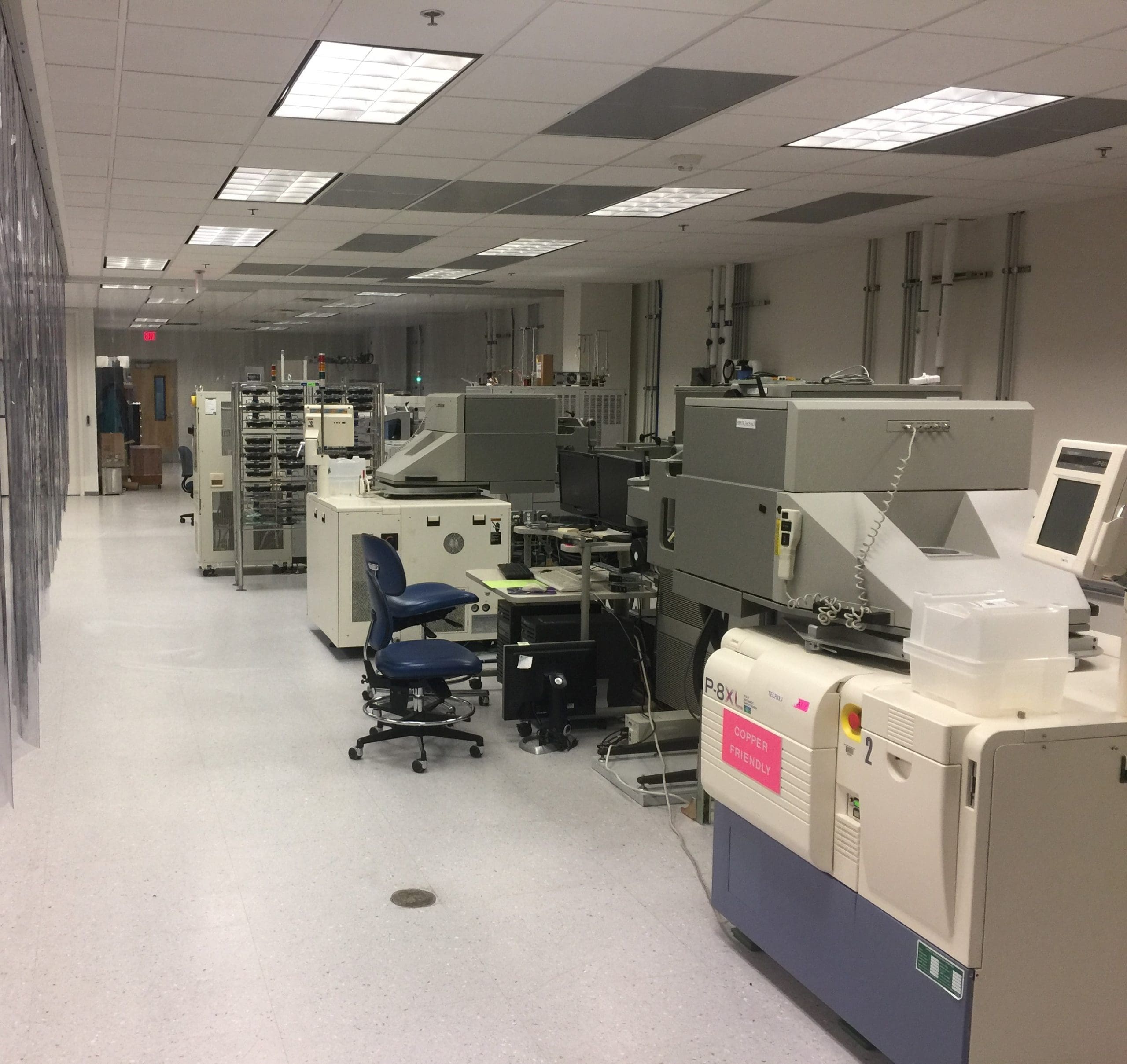 A clean, well-lit laboratory with various machines, computers, and office chairs lined up along the wall, used for research or manufacturing purposes.