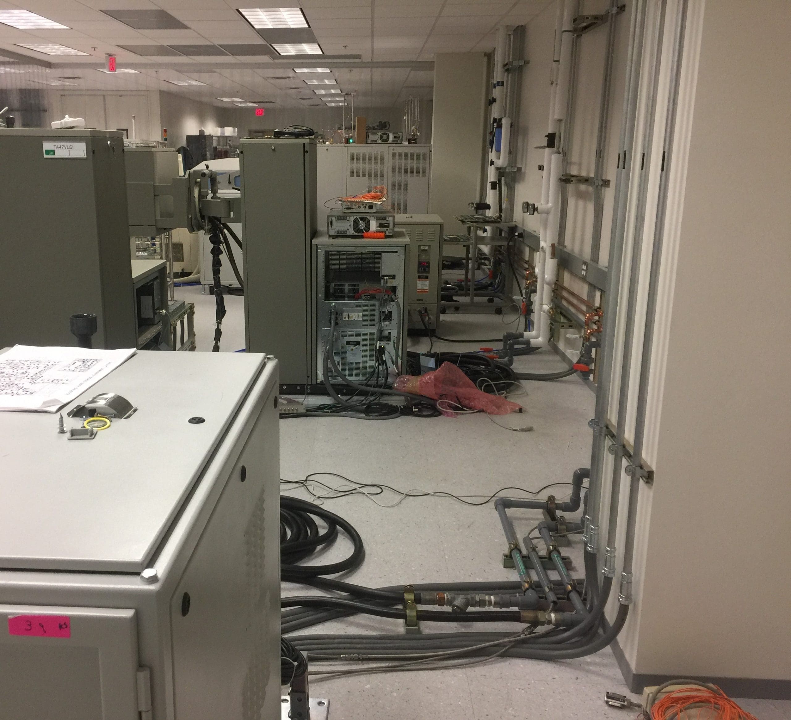Industrial lab room with large electrical equipment, exposed cables, pipes, and various machines positioned against the walls and on the floor.