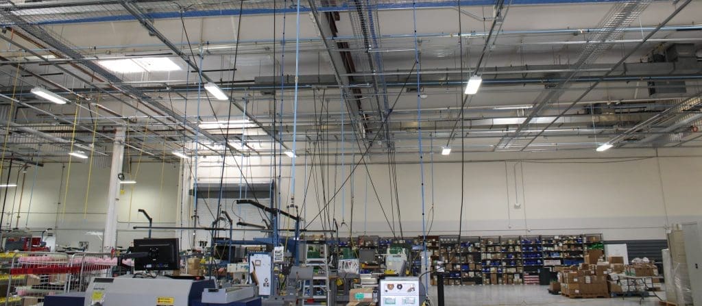 A warehouse interior with shelves of boxes and supplies, machinery, and hanging cables visible against a high ceiling with exposed fixtures.