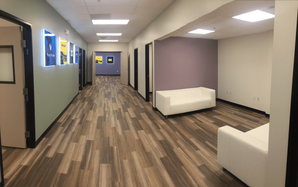 A hallway with wood-patterned flooring, white couches, light green and purple accent walls, and several open doorways leading to offices.