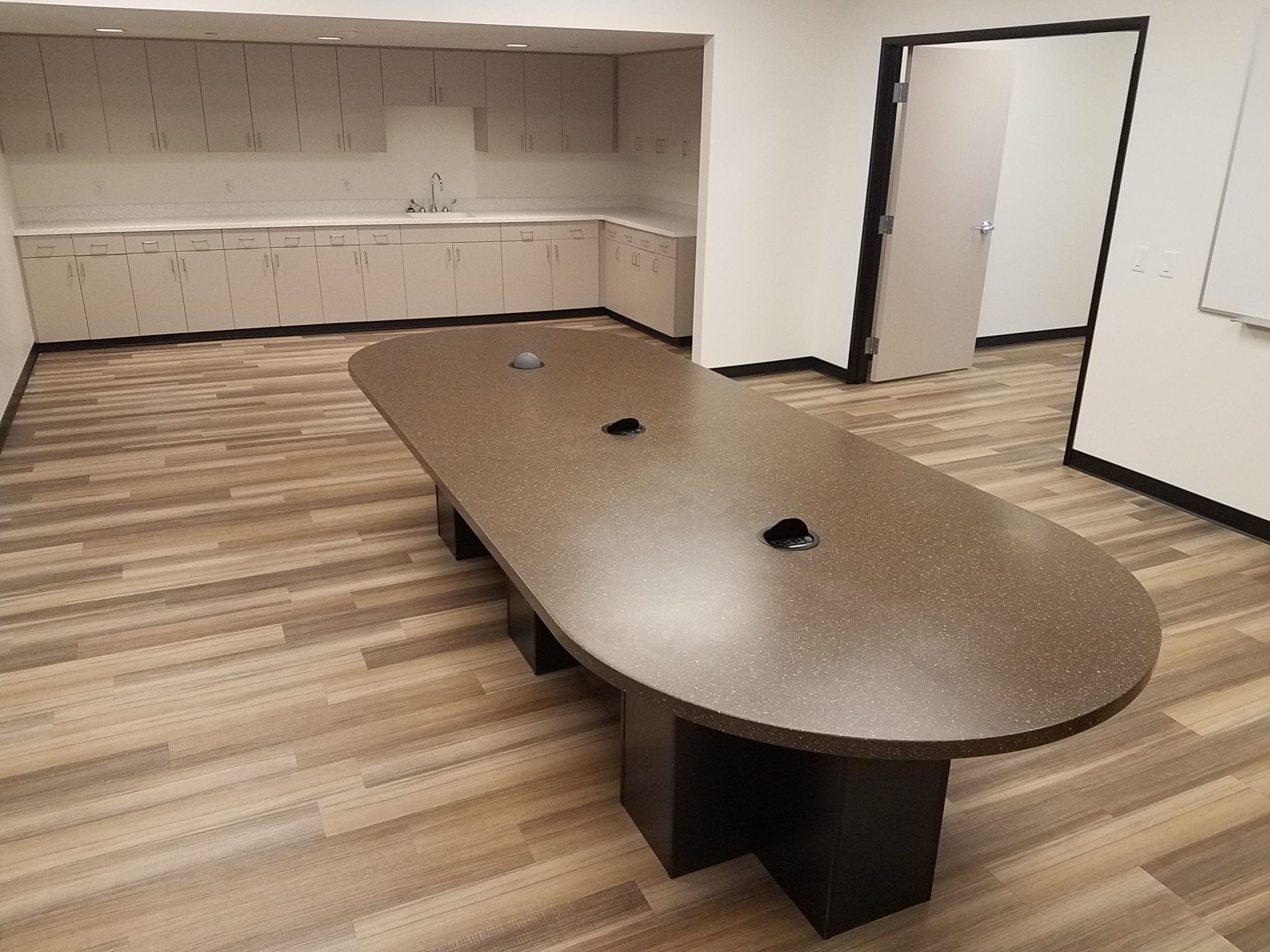 Conference room with a long brown oval table, built-in outlets, wood-patterned flooring, cabinets, countertops, sink, and an open door leading to another room.