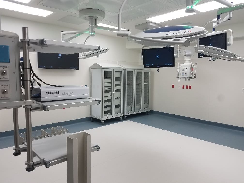 Modern hospital operating room with medical equipment, ceiling-mounted monitors, storage cabinets, and bright overhead lighting.