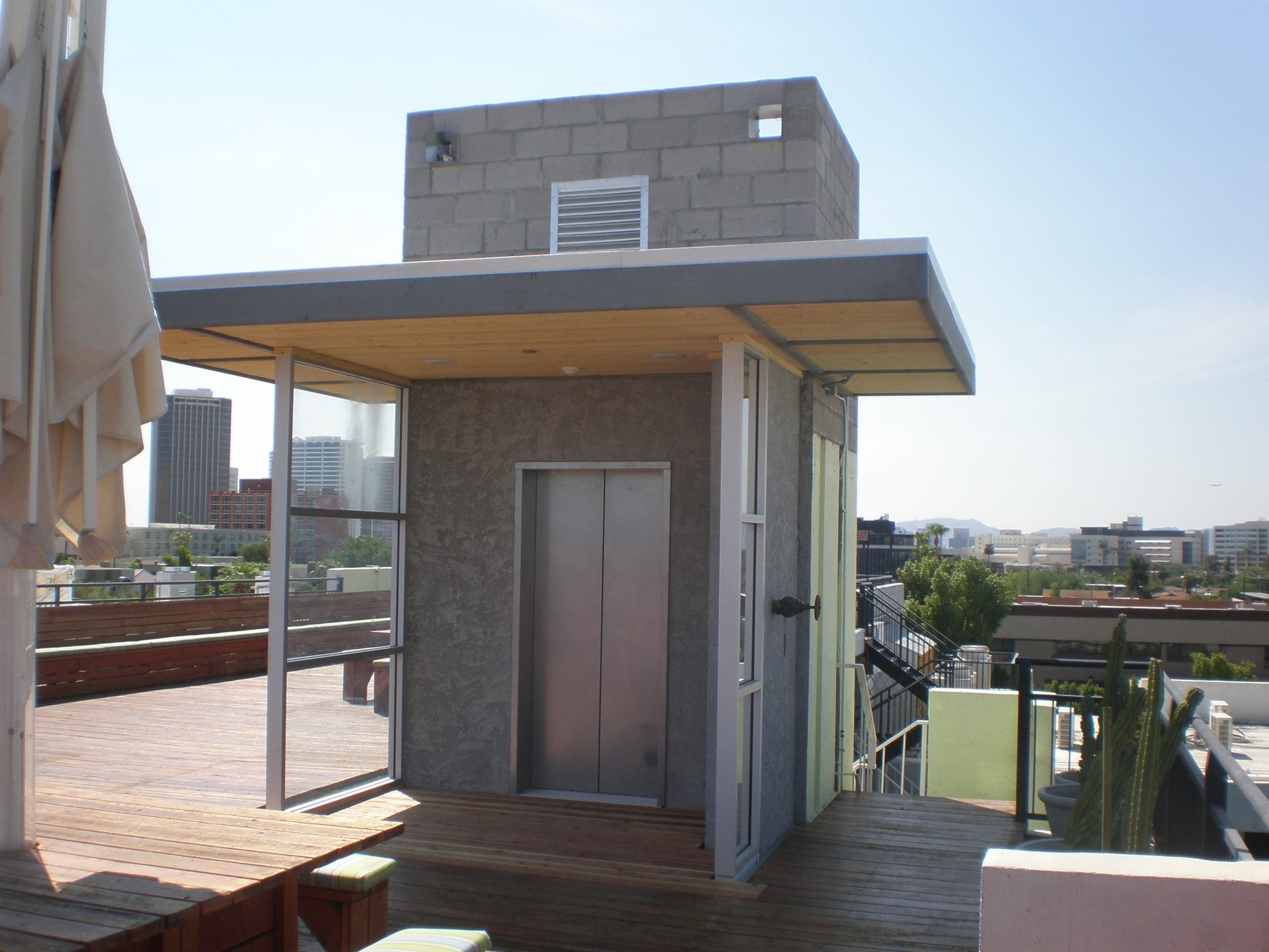 Rooftop area with a modern elevator shaft structure, surrounded by glass railings, overlooking a cityscape on a sunny day.