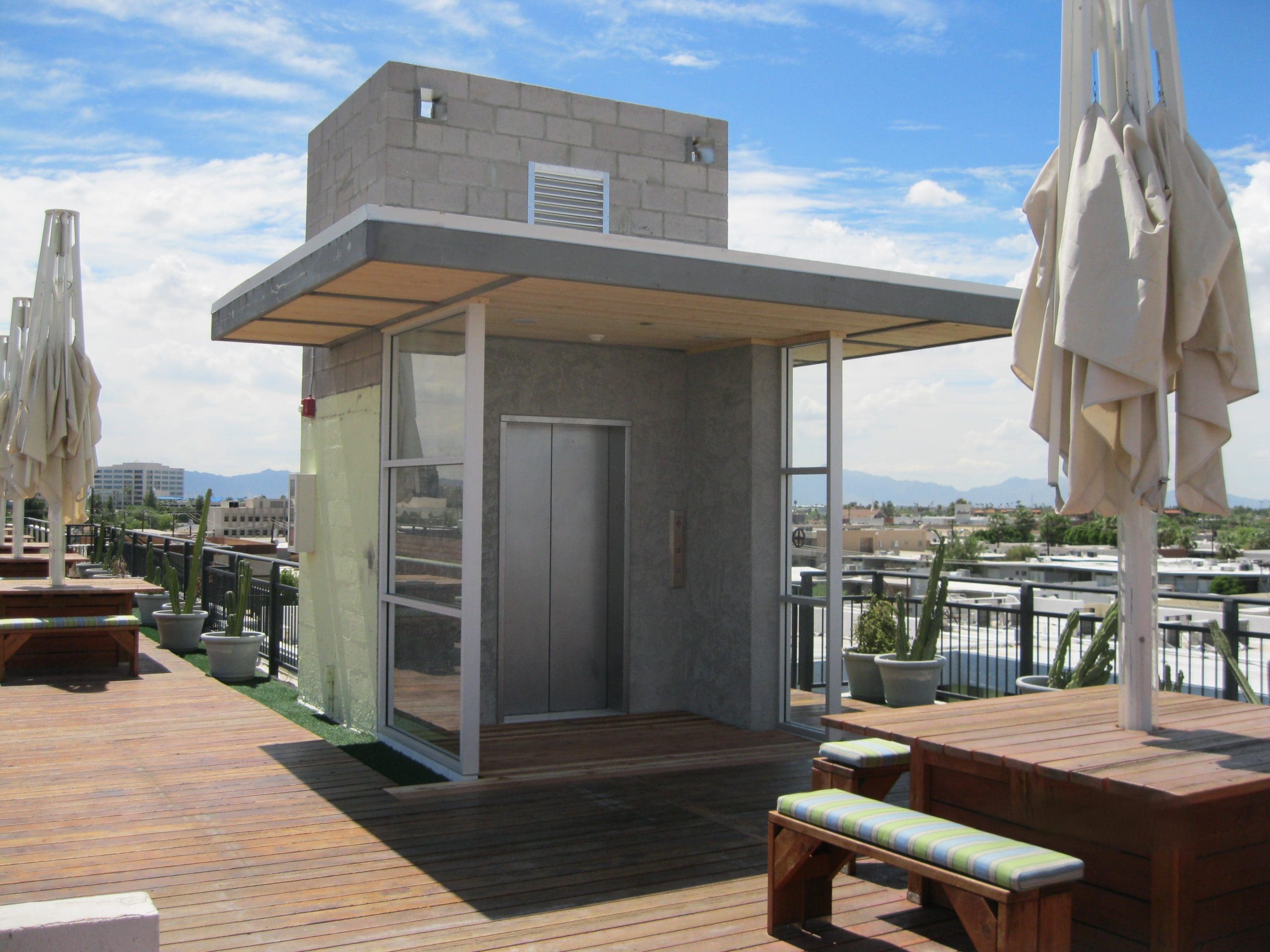 A rooftop patio with wooden benches, closed umbrellas, potted plants, and a modern elevator structure in the center under a partly cloudy sky.