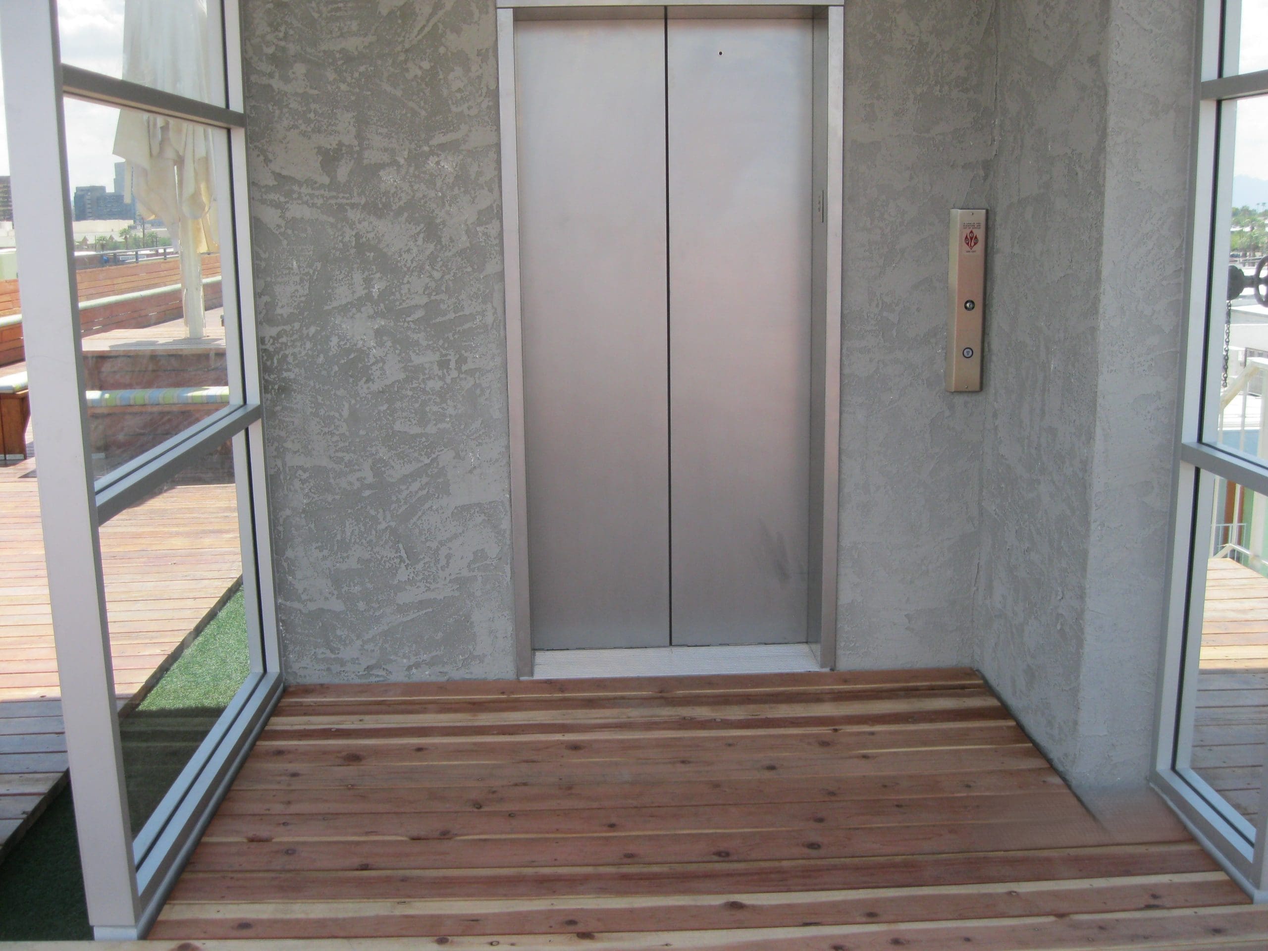 A closed stainless steel elevator door in a small alcove with wooden flooring and textured gray walls. Elevator buttons are mounted on the right side.