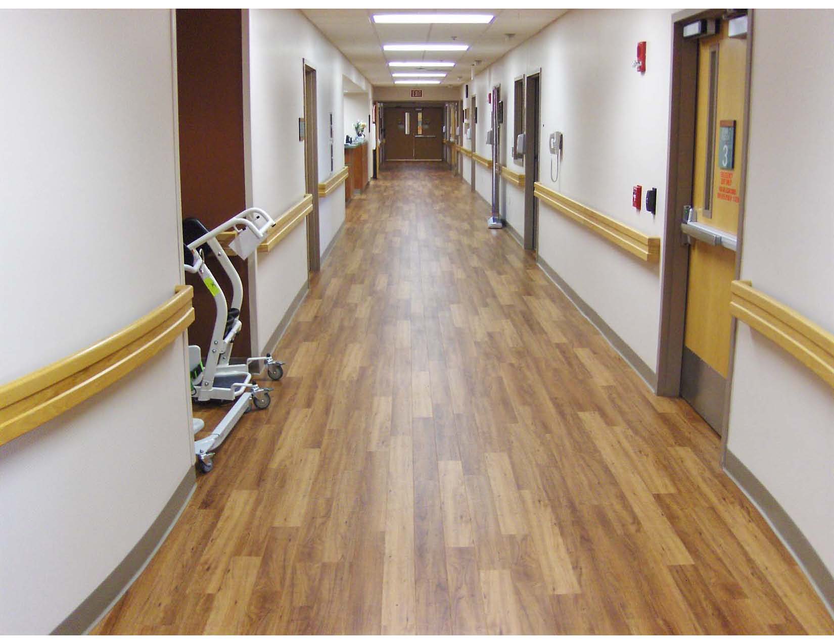 A clean, empty hospital hallway with wood-patterned flooring, beige walls, yellow handrails, and medical equipment on the left side. Doors and ceiling lights line the corridor.