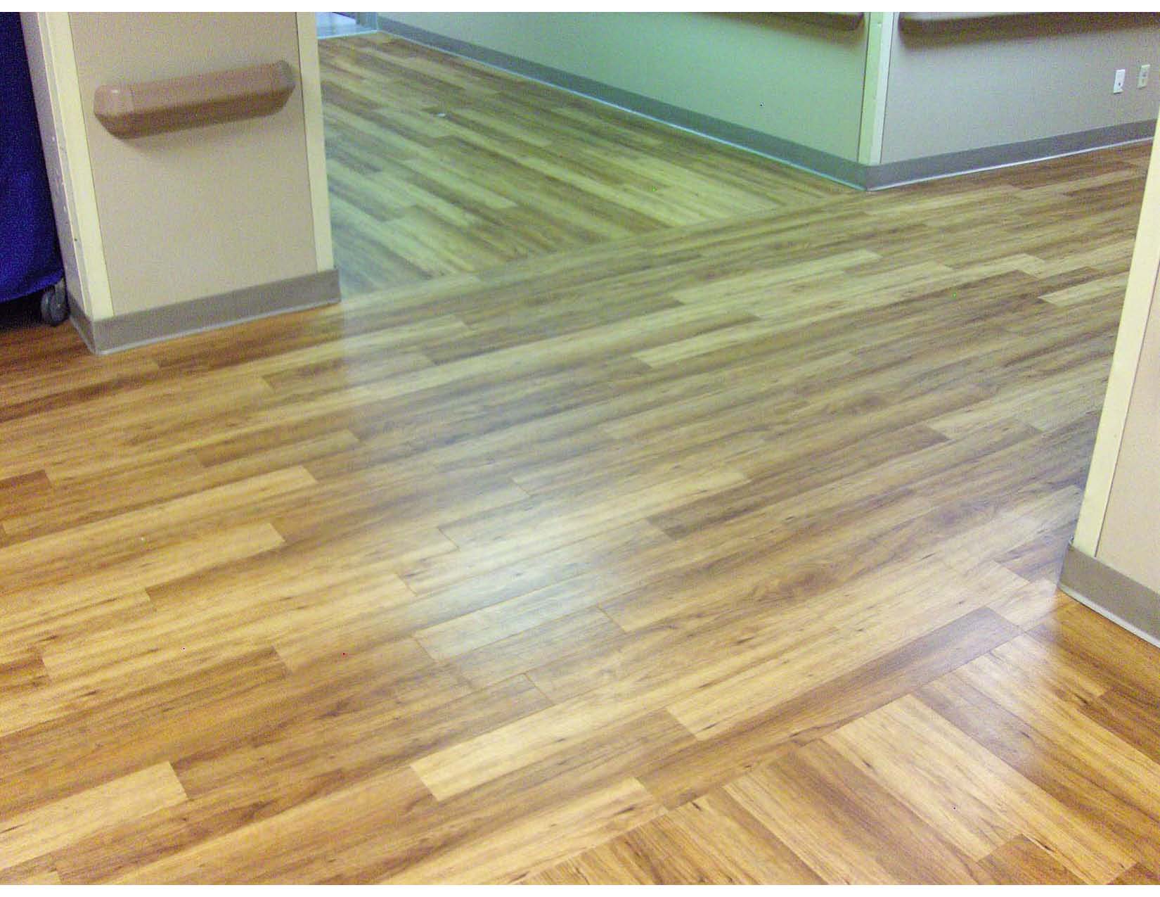 A hallway with smooth, light brown wood-patterned flooring, beige walls, and baseboards, extending into another room with the same flooring.
