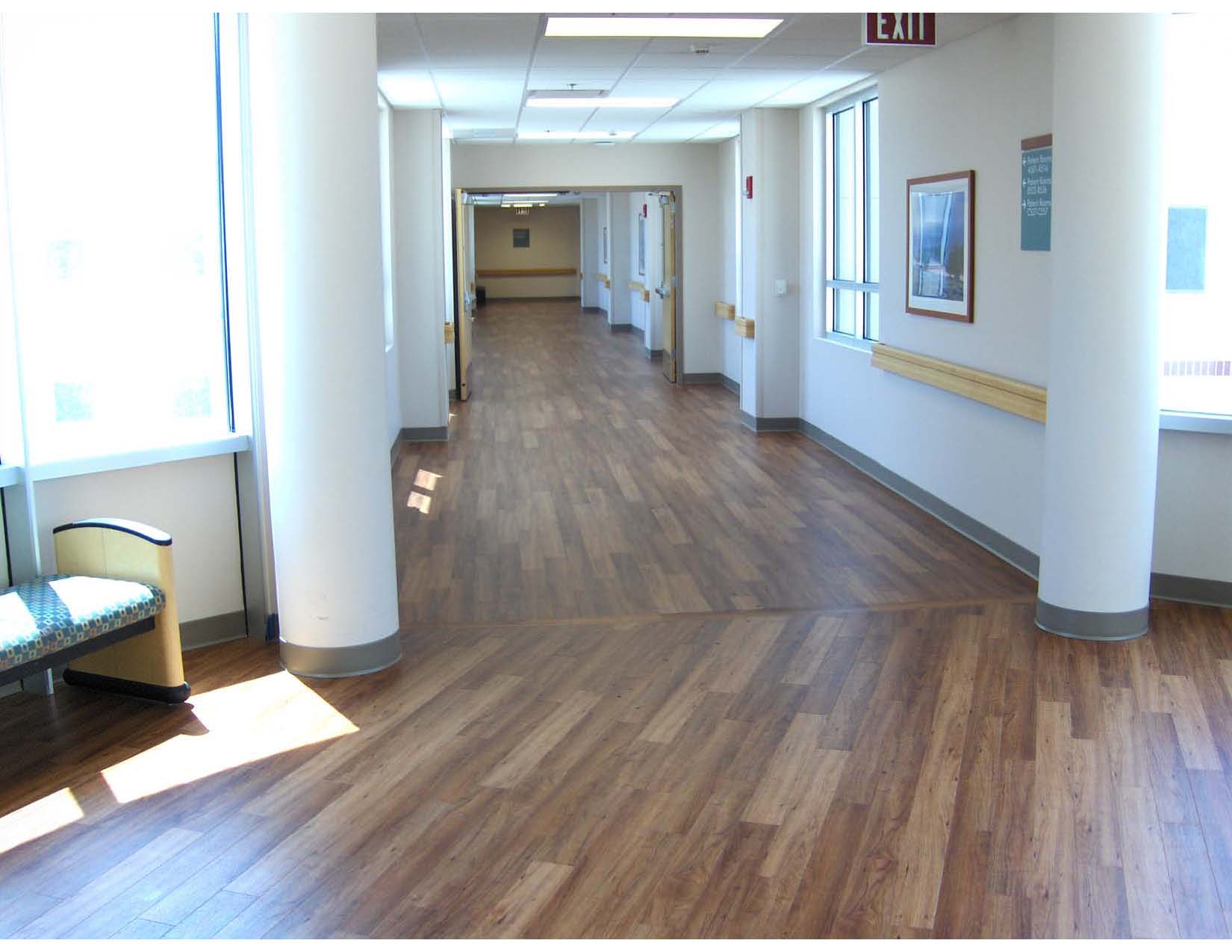 A brightly lit hallway in a building with wood-patterned flooring, white walls, handrails, a bench, and framed artwork on the walls.
