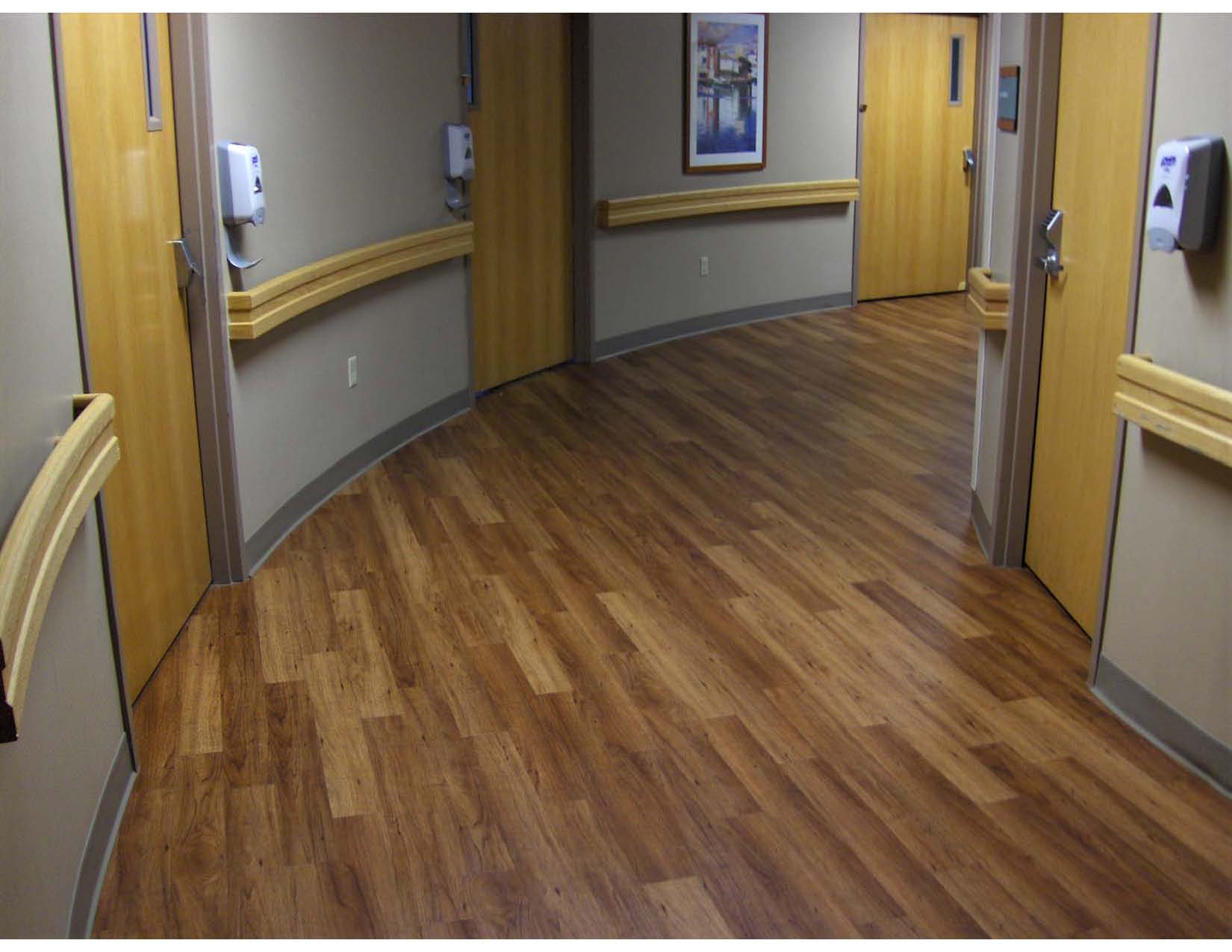 A hospital hallway with wood-look flooring, beige walls, yellow handrails, closed wooden doors, wall-mounted hand sanitizers, and a framed picture on the wall.