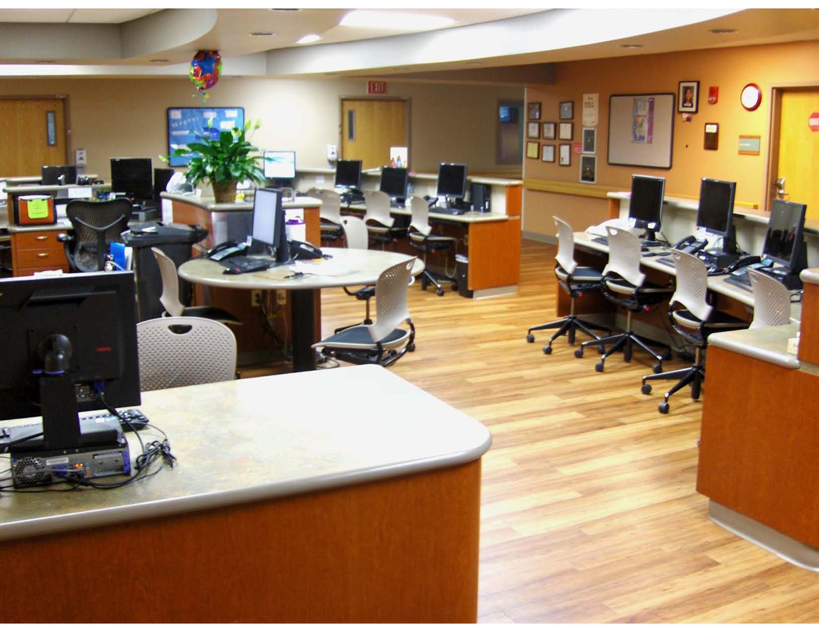 A hospital or medical office workstation area with multiple desks, computers, and chairs arranged in a clean, organized setting on a wood-style floor.