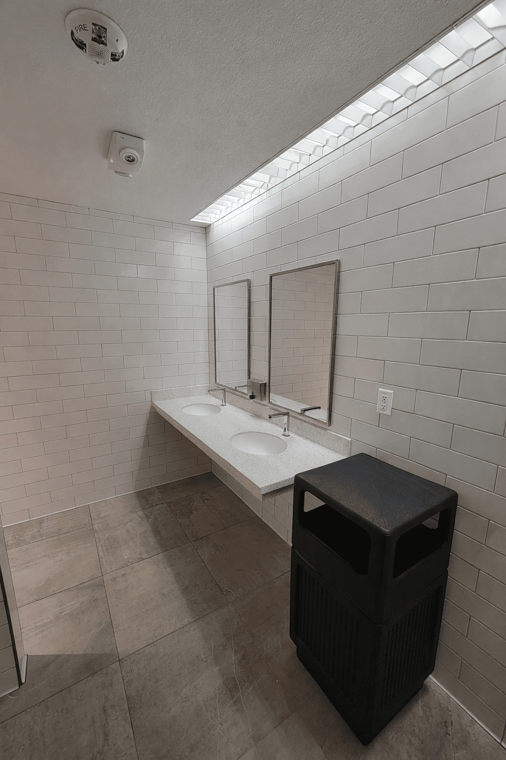 A public restroom with two sinks, two mirrors, white tiled walls, beige tile flooring, and a large black trash bin.
