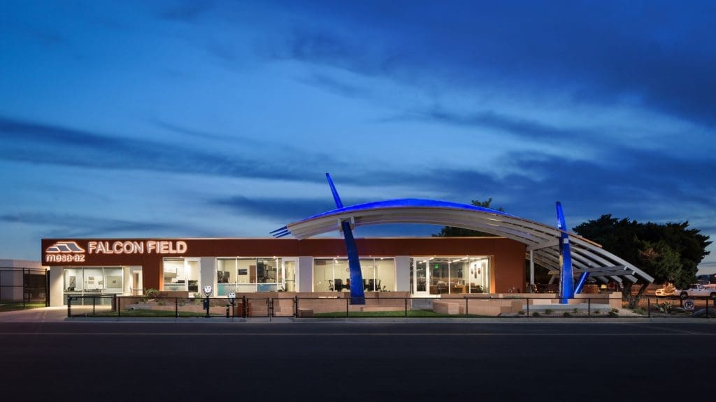 Modern building labeled “FALCON FIELD” with Mazda logo, large windows, and a curved roof structure, illuminated by lights at dusk.