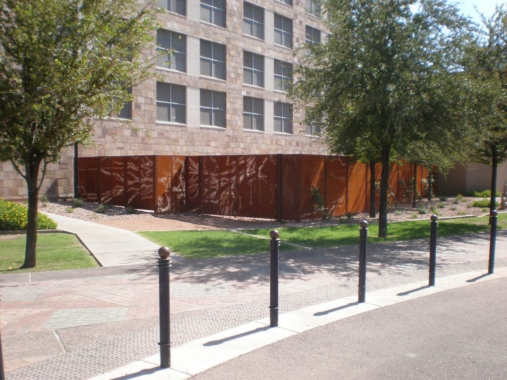 A modern building with stone walls, trees, a sidewalk, and a rust-colored metal fence. Black bollards line the edge of the street in the foreground.