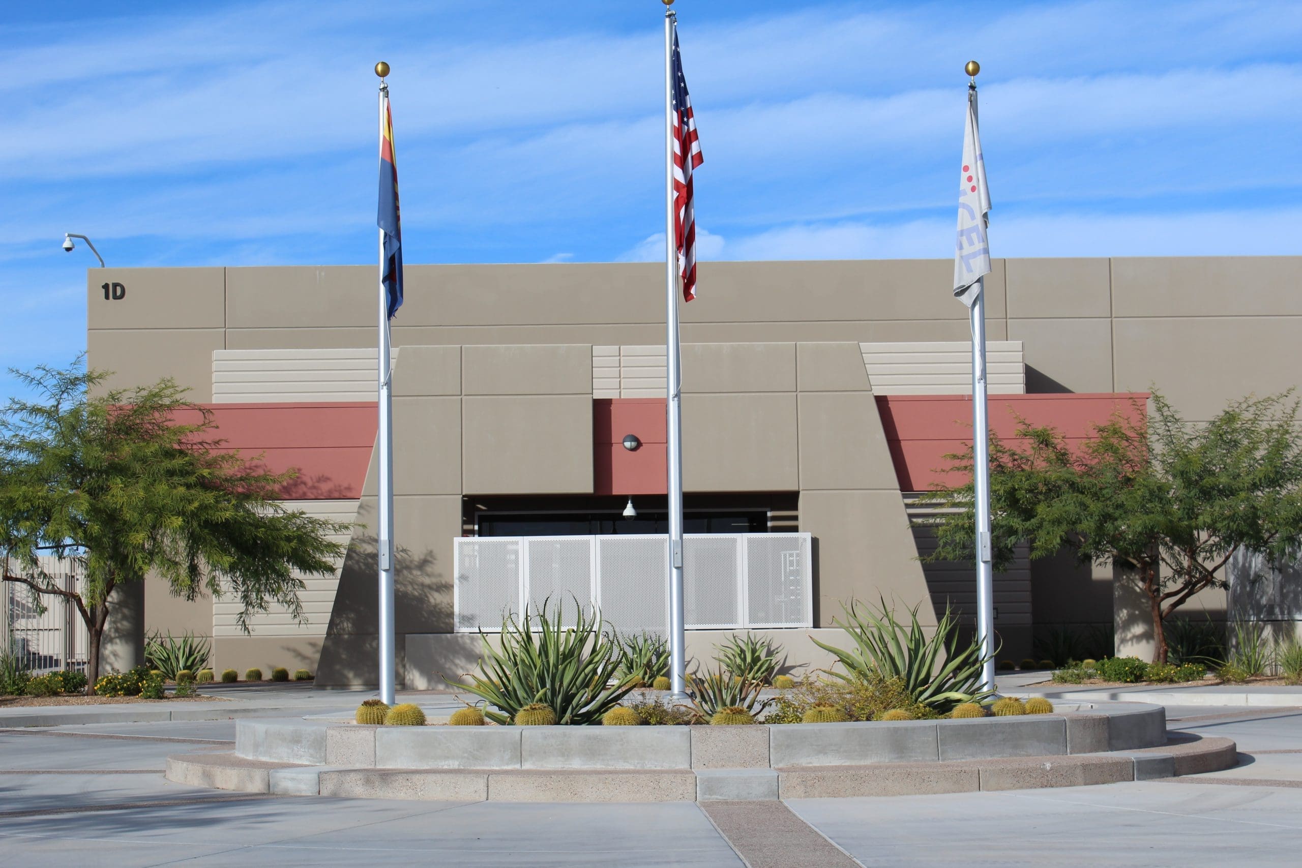A beige building with three flagpoles in front, displaying the Arizona state flag, the U.S. flag, and a white flag, with desert landscaping and a blue sky overhead.