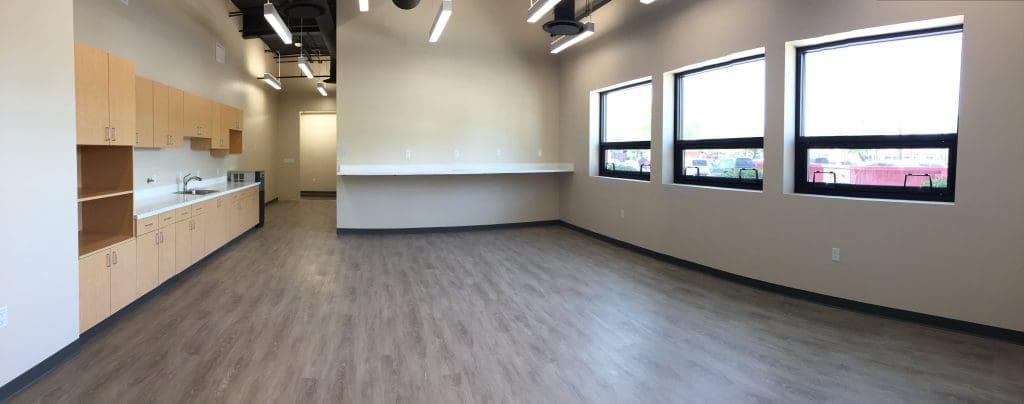 A spacious, unfurnished room with wood-style flooring, large windows, ceiling lights, and a kitchenette with light wood cabinets along one wall.