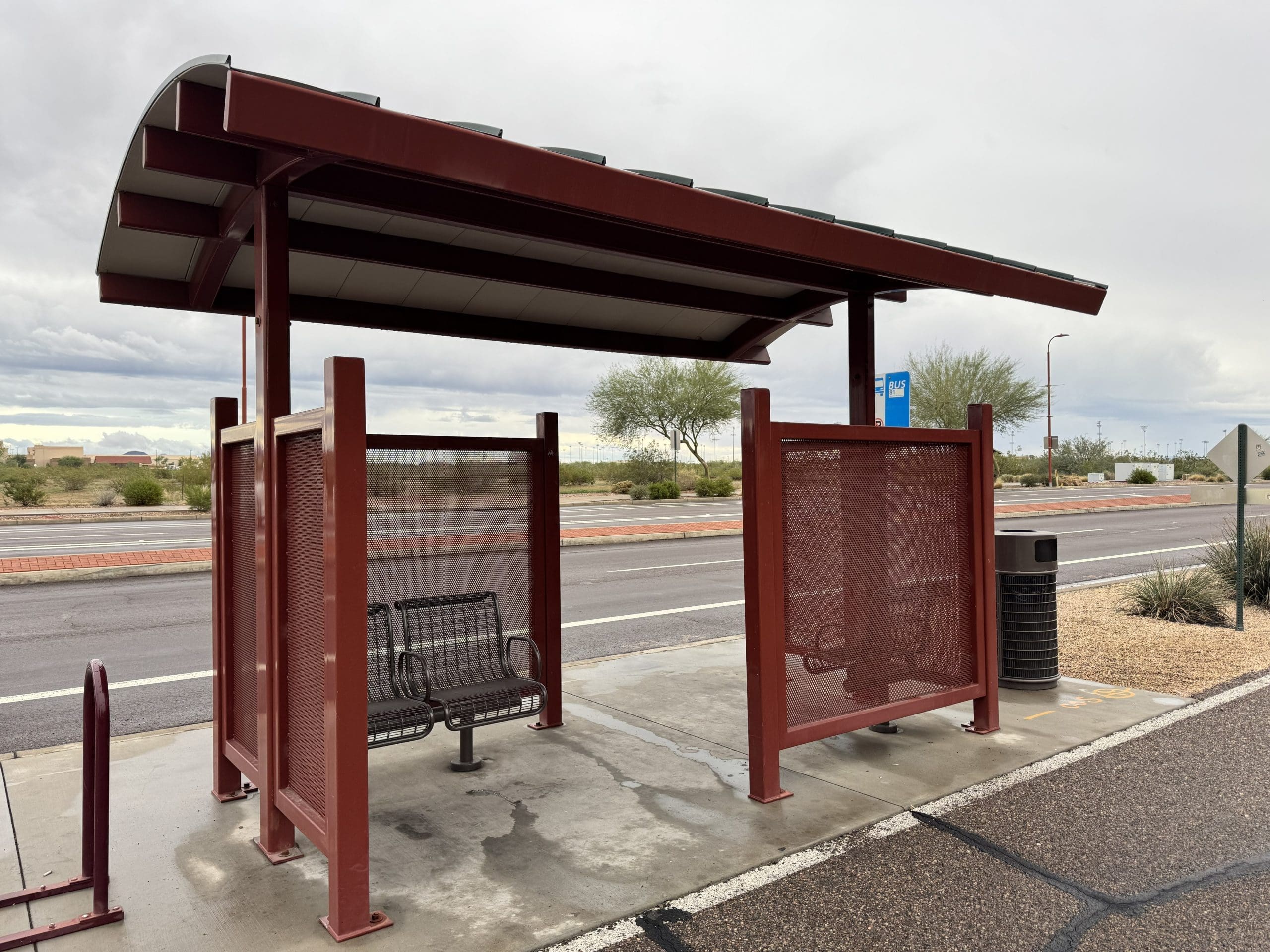 A covered bus stop with metal benches, a trash can, and mesh side panels stands beside a road on a cloudy day in a desert area.