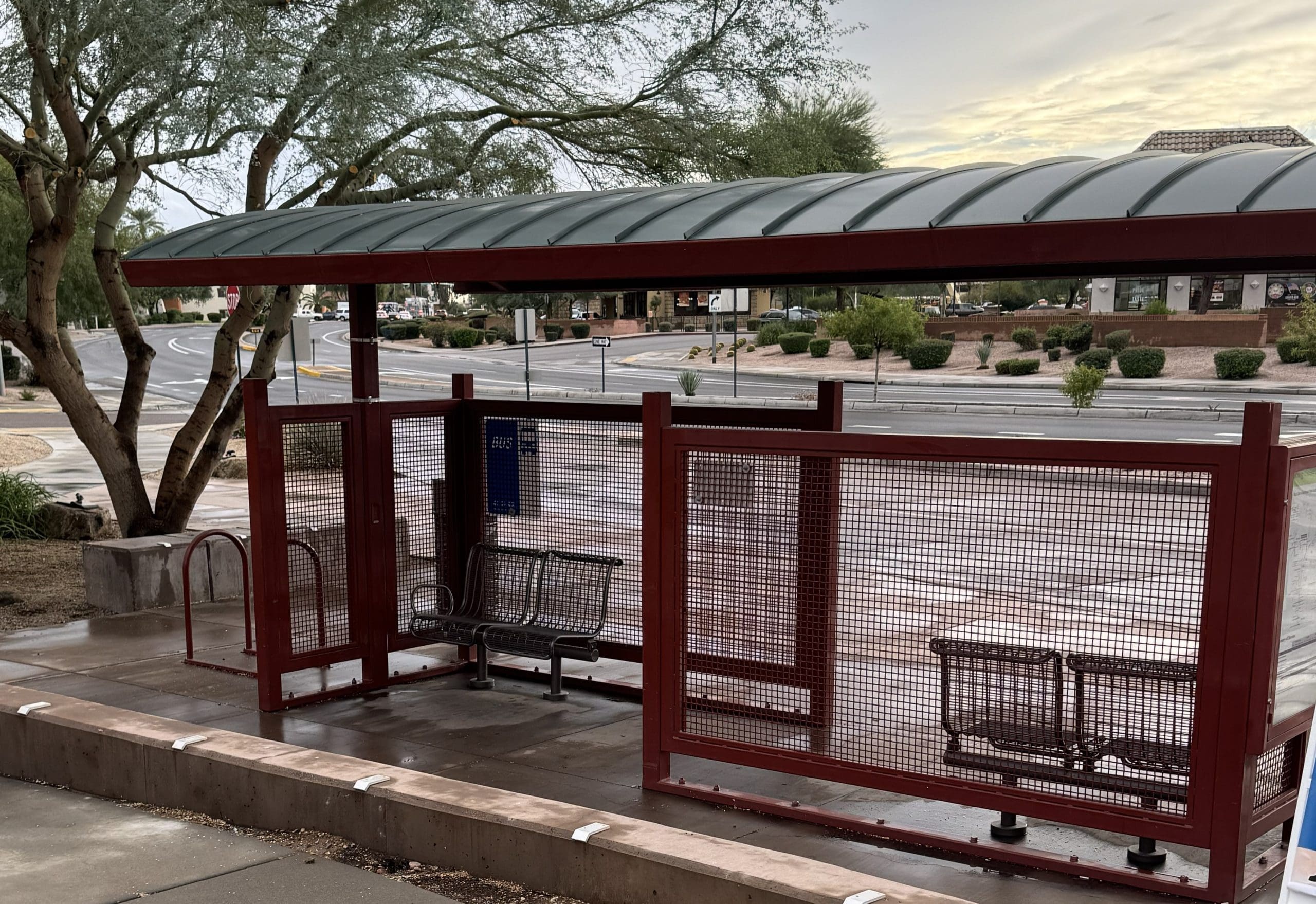 A covered bus stop with metal benches and a bike rack sits empty on a wet sidewalk near a suburban street, with trees and buildings in the background.