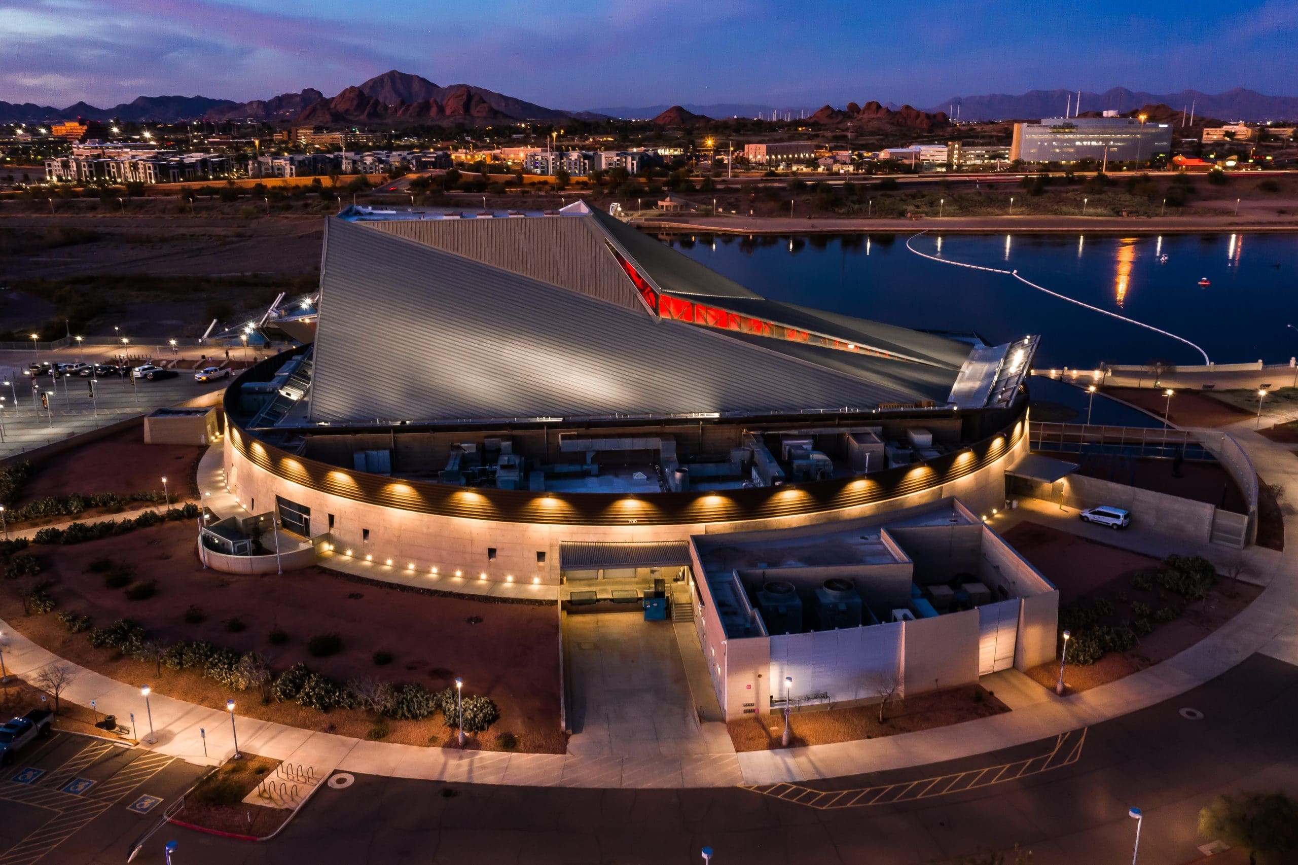 Aerial view of a modern, circular building with angled roof, surrounded by a lit pathway, water, and cityscape at dusk.