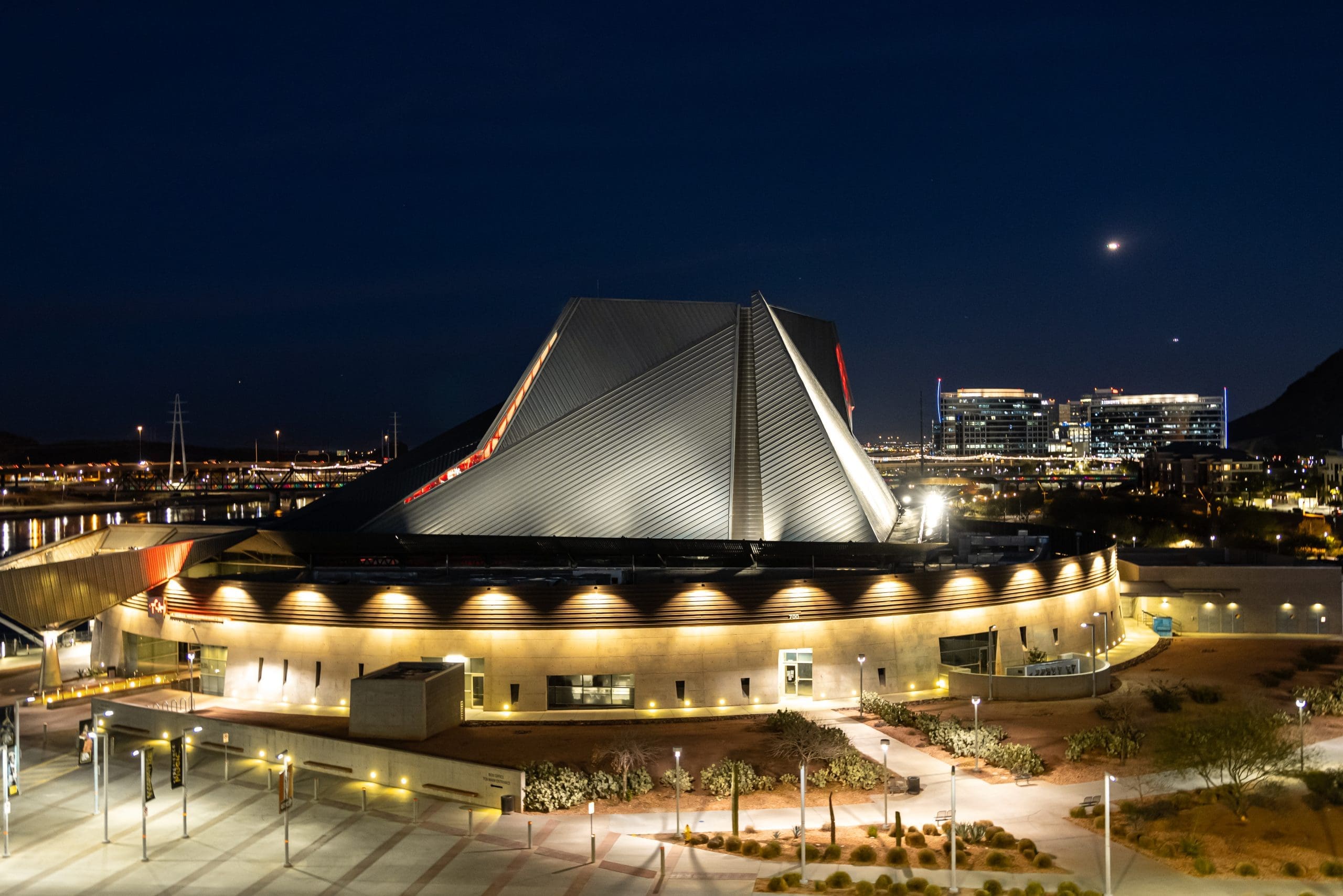 A modern, angular building is illuminated at night, with city lights and office buildings in the background under a dark sky.