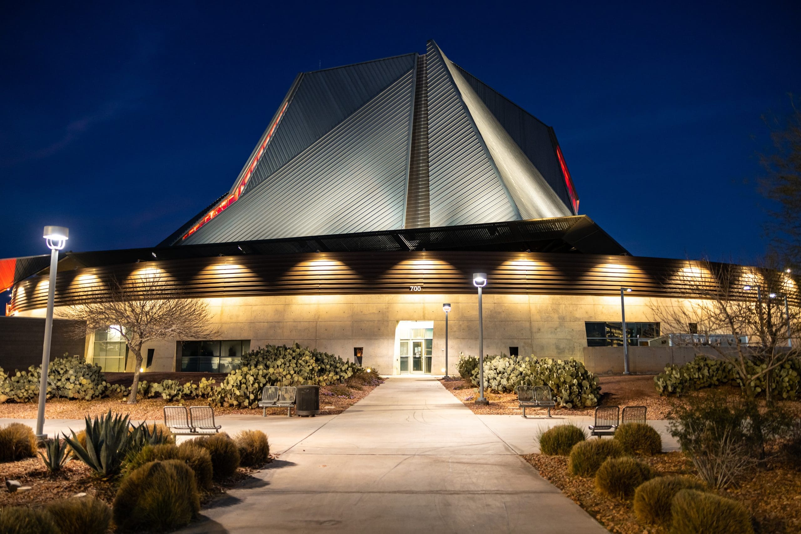 A modern, angular building with illuminated exterior lights and desert landscaping is pictured at night, with a clear pathway leading to the entrance.