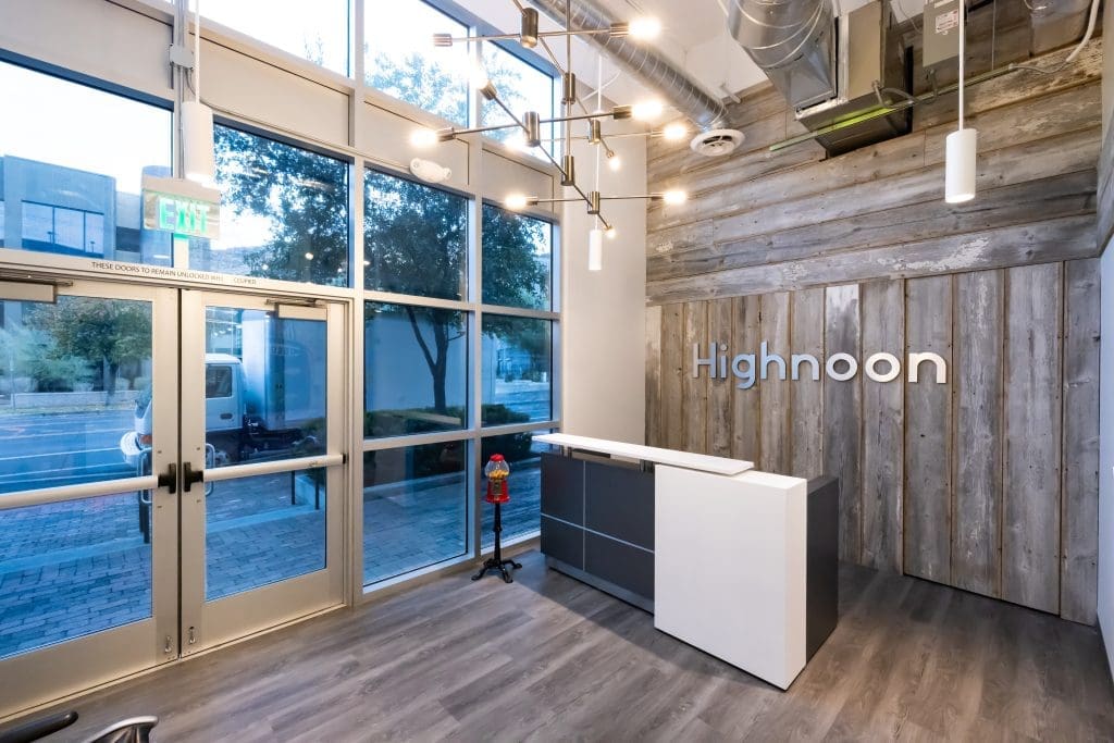 Reception area with wood-paneled wall, "Highnoon" sign, modern desk, large windows, and an exit door leading outside.