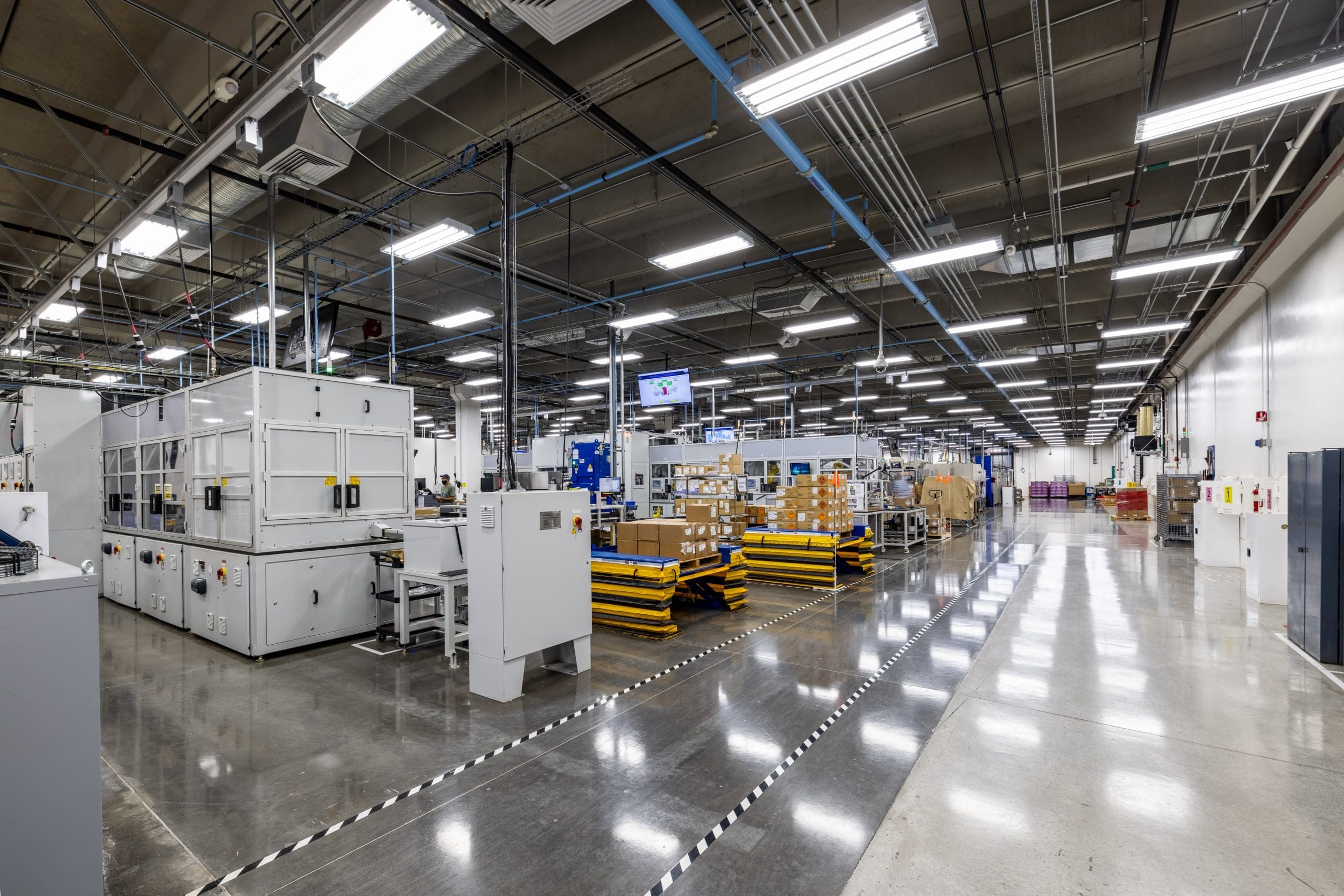 A clean, modern industrial facility with automated machinery, stacked cardboard boxes on pallets, and bright overhead lighting.