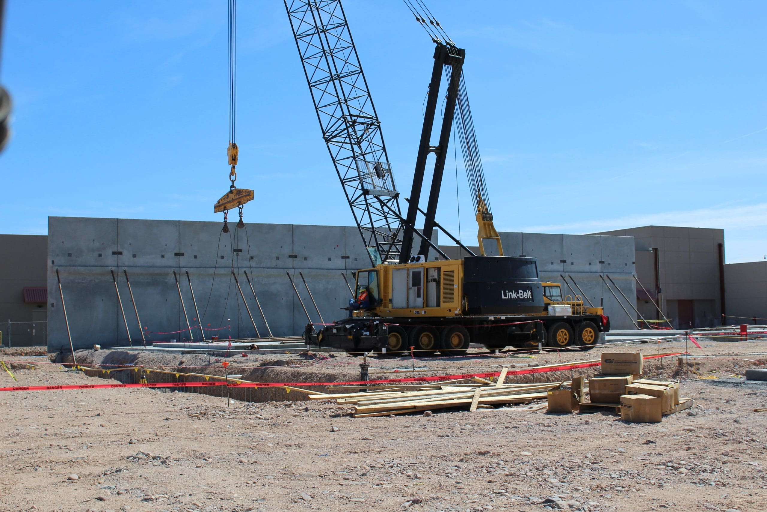 A crane lifts a concrete panel at a construction site with workers, scattered lumber, and safety barriers visible under a clear blue sky.