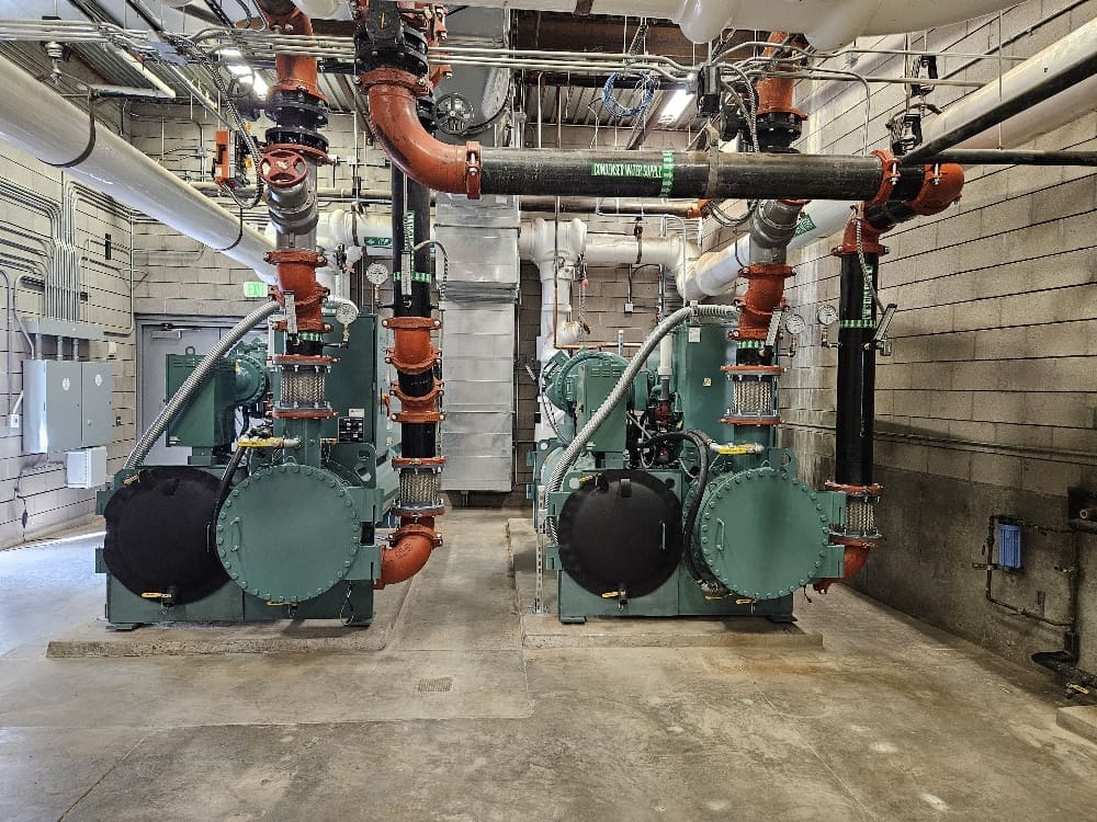 Two large industrial machines with pipes and valves are installed in a concrete-floored utility room with brick walls and exposed ceiling infrastructure.